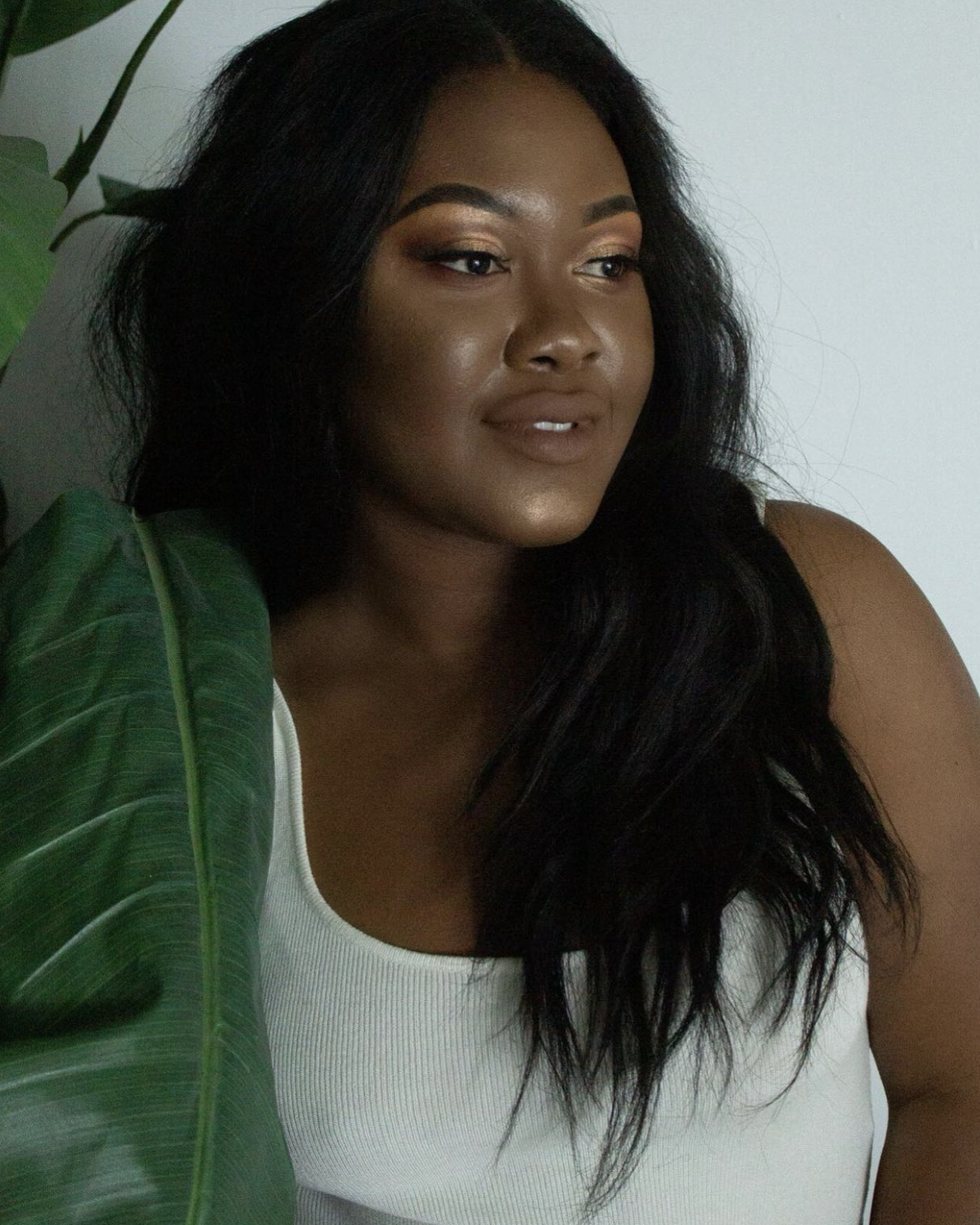 A woman with long black wavy hair, wearing a white tank top, is next to a green large leaf plant against a white wall.