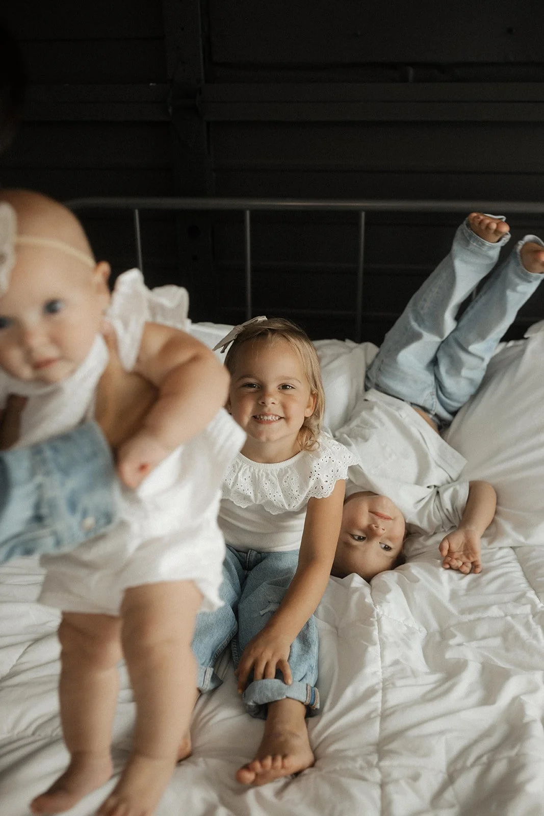 Three children playing on a bed, smiling and lying down in a cozy bedroom with a dark headboard.