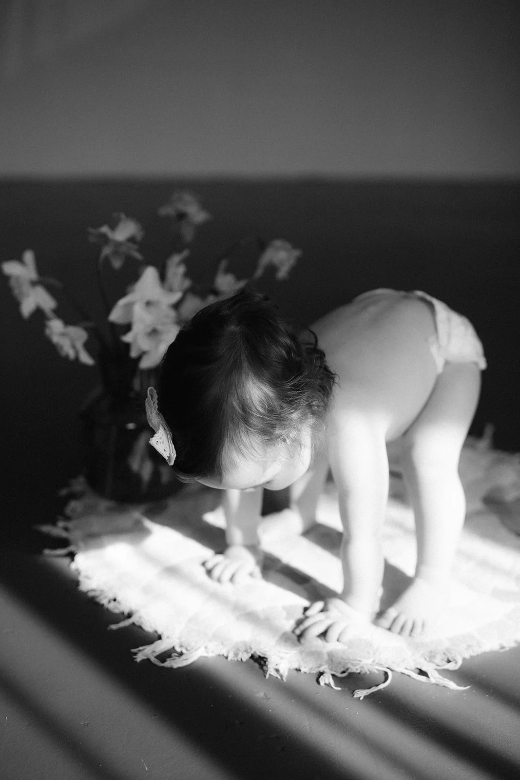 A young girl with a butterfly hair clip kneeling on a rug, with a vase of flowers behind her, in black and white.