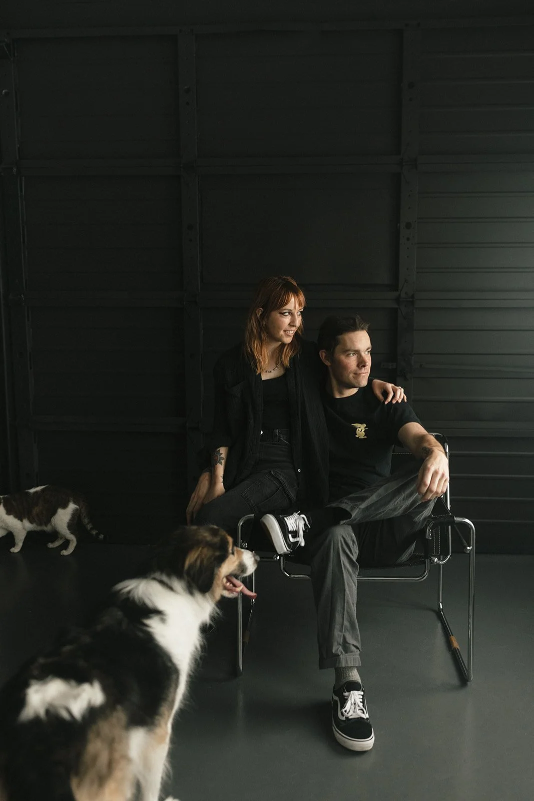 A woman and a man sit together, with the woman's arm around the man's shoulders, inside a dark, industrial space. There are two dogs, one in the foreground and one in the background.