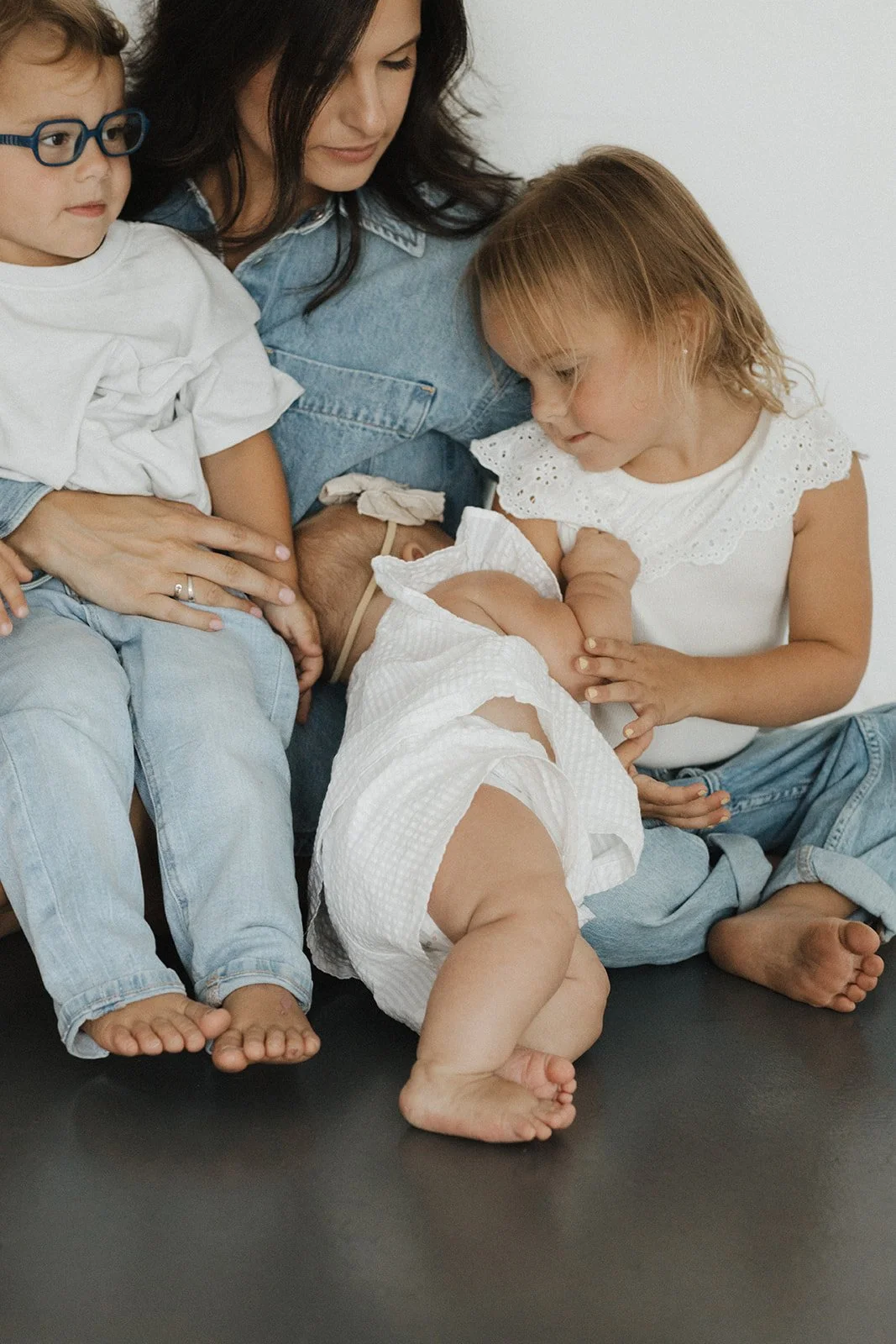 A woman and three children, with one baby, sit closely together, with the woman breastfeeding the baby. The children are engaging with each other and the baby, in a cozy white and denim setting.