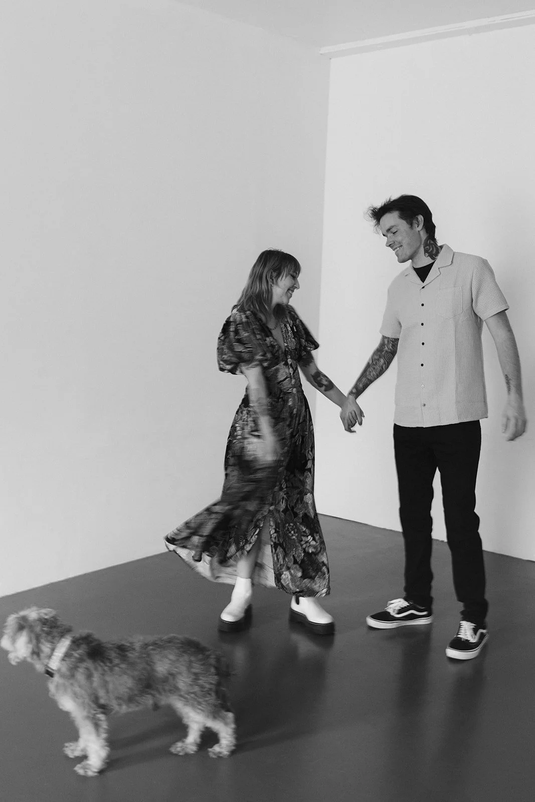 A smiling woman and a smiling man holding hands and dancing together in a room with a dog walking in front of them.