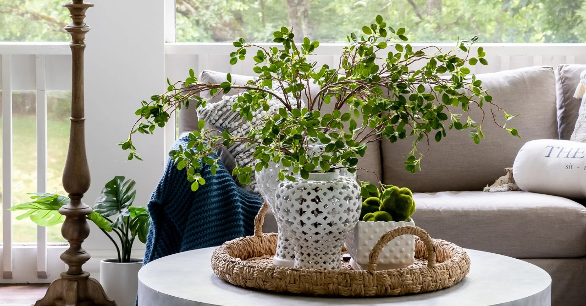 Bright summer home living room featuring lush green plants, a round concrete coffee table, and natural light flooding through large windows.