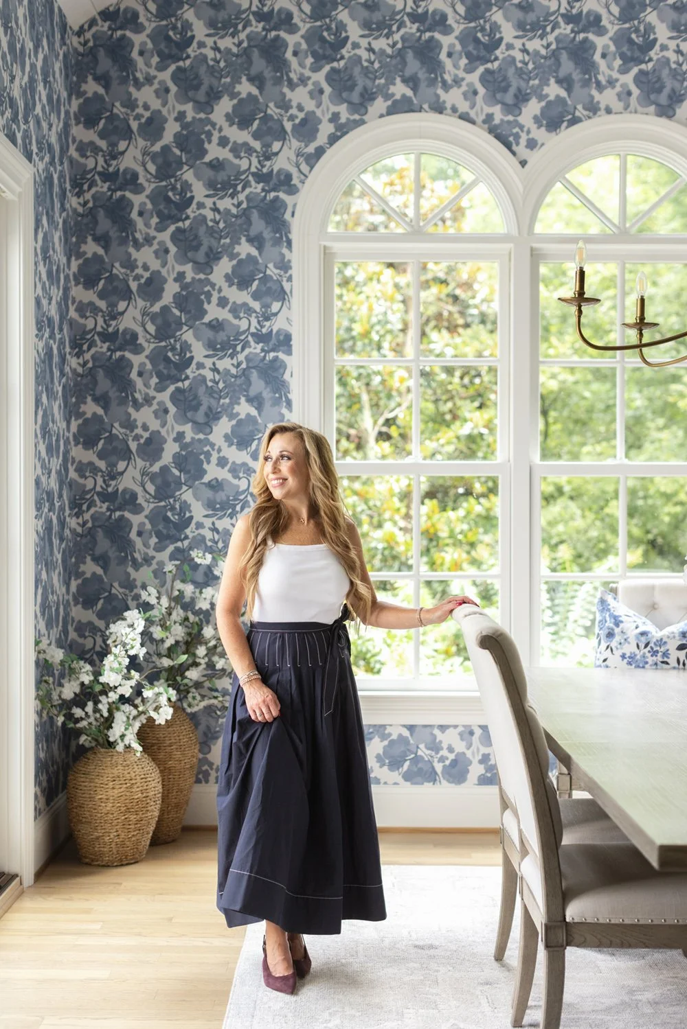 Interior designer standing by arched windows in a dining room with blue floral wallpaper and a rustic farmhouse table