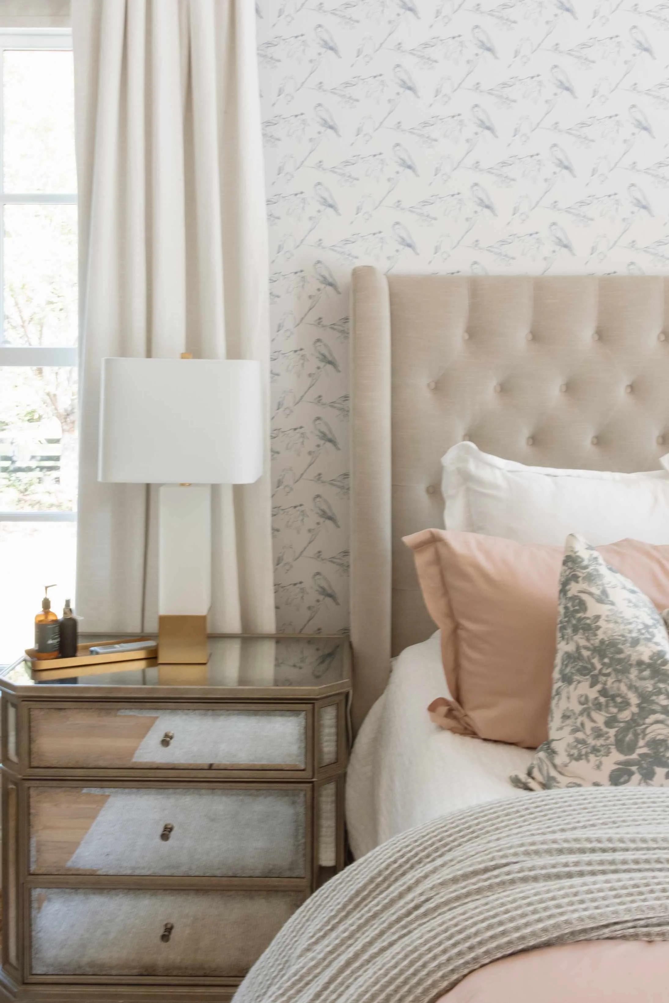 Interior decorating tips for bedroom with bird print wallpaper accent wall, tufted headboard, mirrored nightstand, and blush pink pillows.