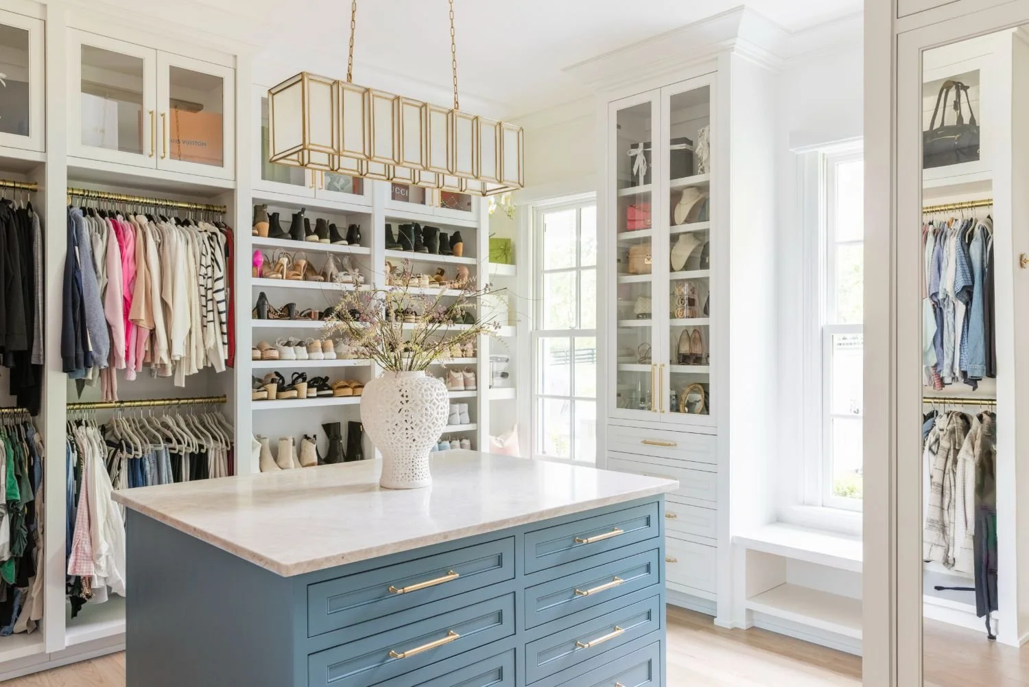 A spacious, luxurious, and modern walk-in closet with rows of hung up clothes, women's shoes, and women's accessories. Florals in a white vase sit on a blue closet island, which is located in the center.
