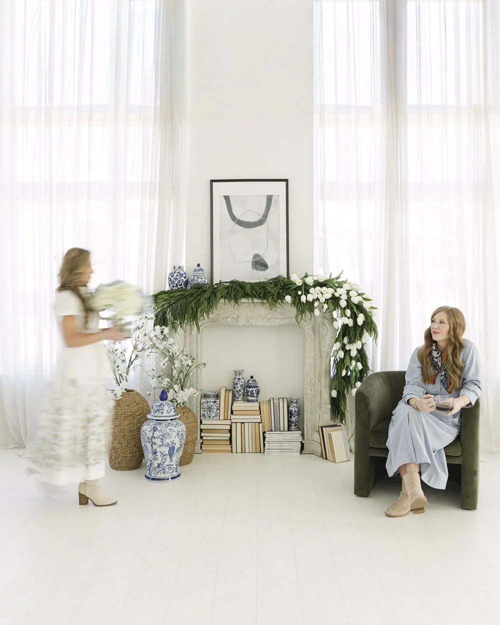 Two interior designers in a bright studio with a garland-draped mantel, blue-and-white ginger jars, and a green velvet chair