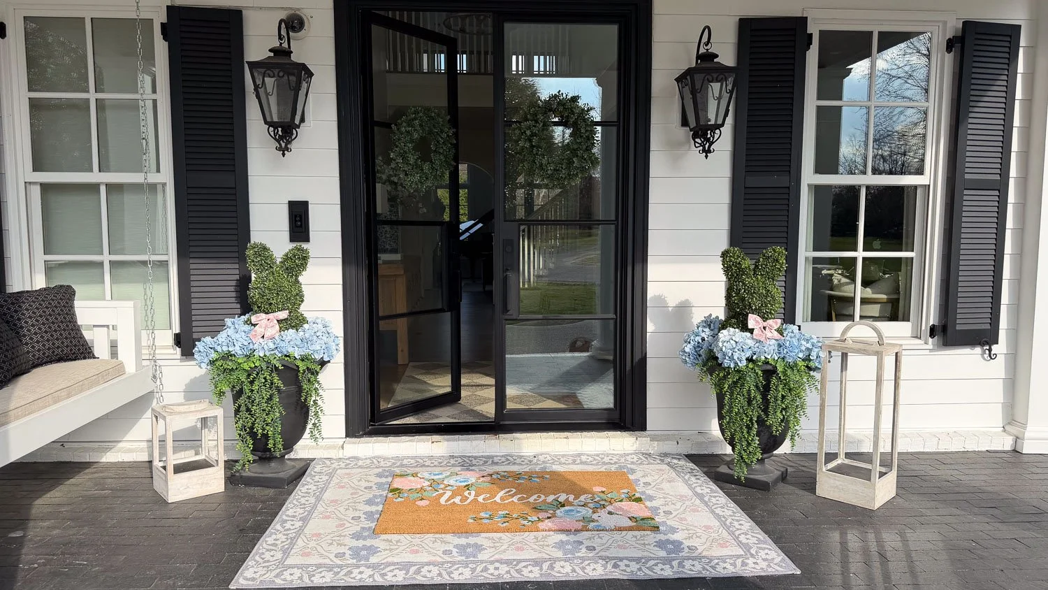 A front entryway featuring spring decor with blue hydrangeas, a white lantern, and a colorful floral doormat.