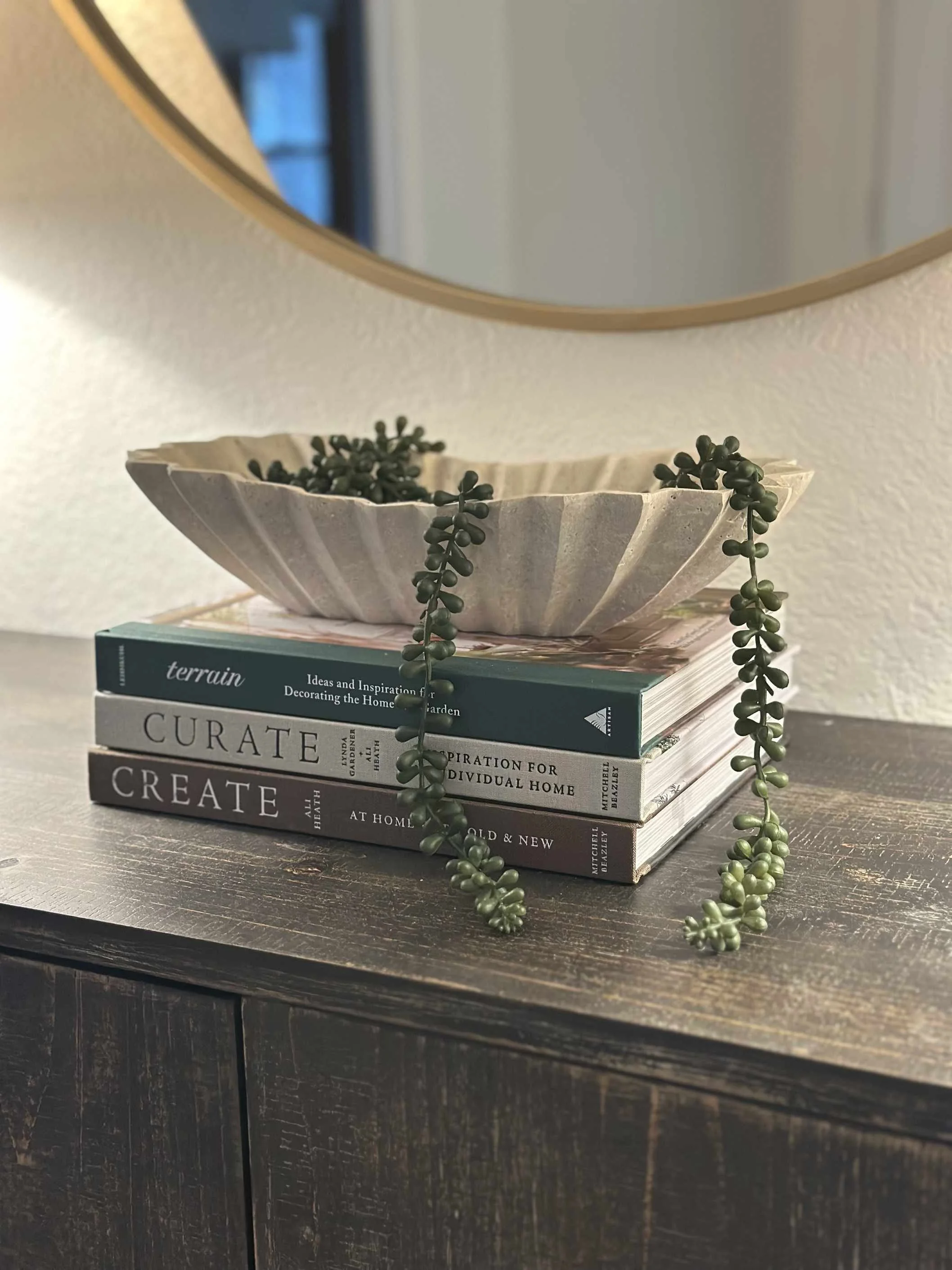 Interior decorating tips for table styling with stacked design books, ceramic bowl with trailing succulent, and round gold mirror above.