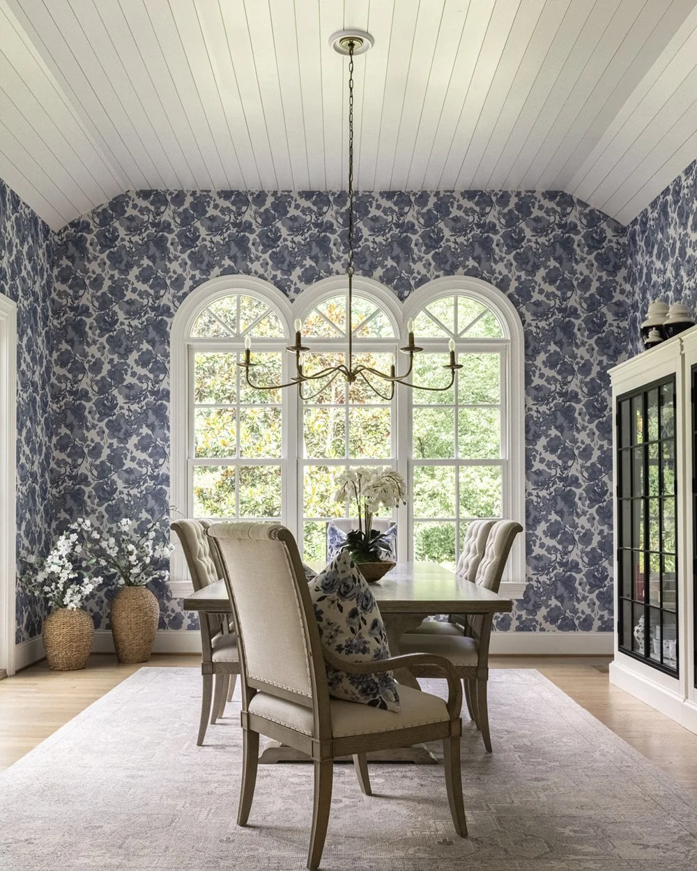 Luxury dining room with blue watercolor floral wallpaper, triple arched windows, a brass chandelier, and upholstered dining chairs.