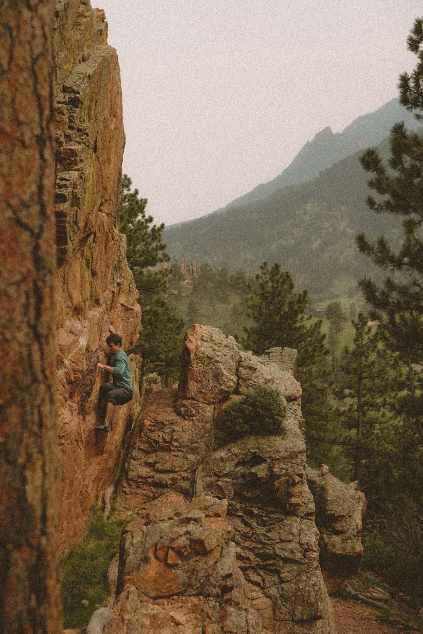 A person rocks climbing on a red rocky cliff with mountain and forest landscape in the background.