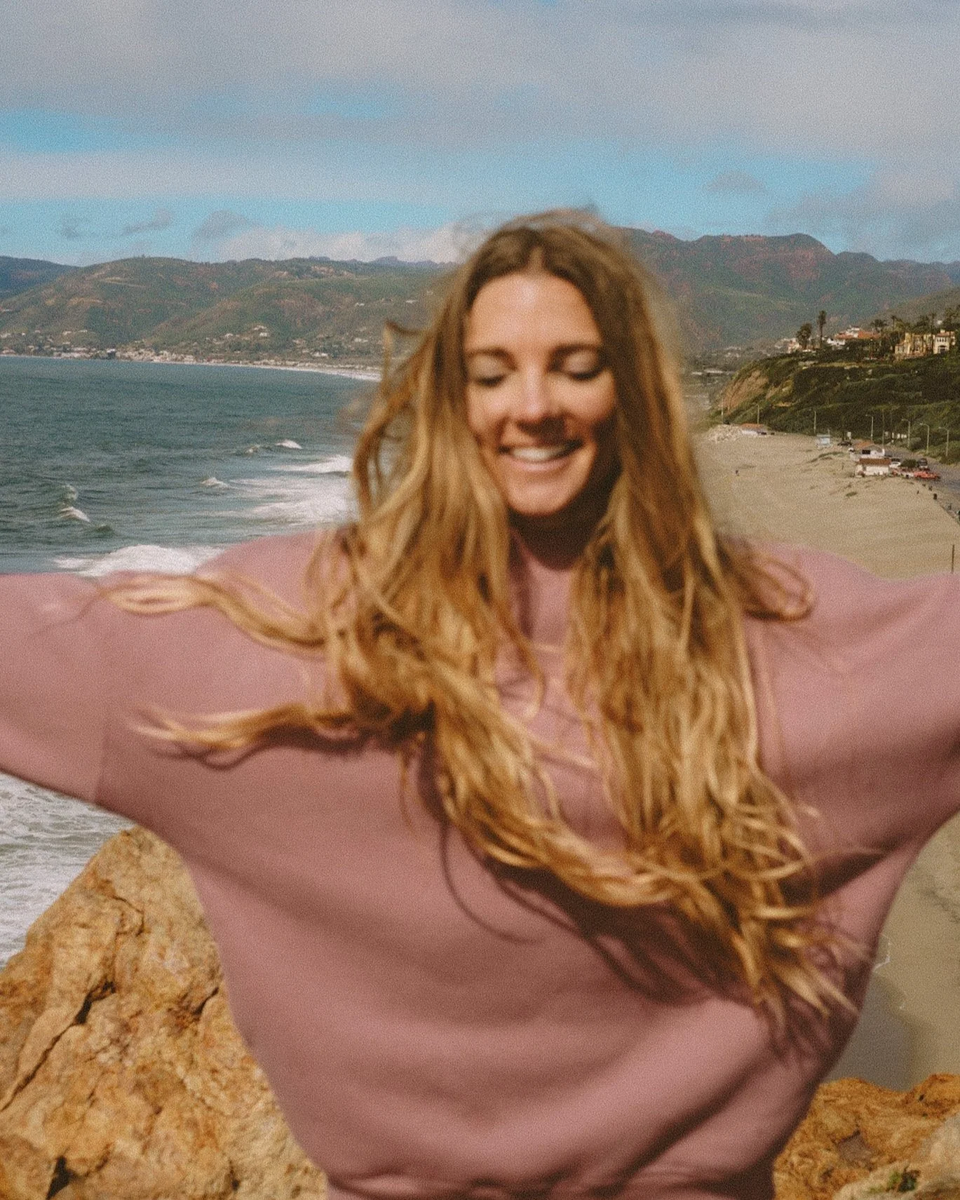 A woman with long, wavy, reddish-brown hair smiling with arms outstretched on a beach, with ocean waves, sandy shore, cliffs, and mountains in the background.