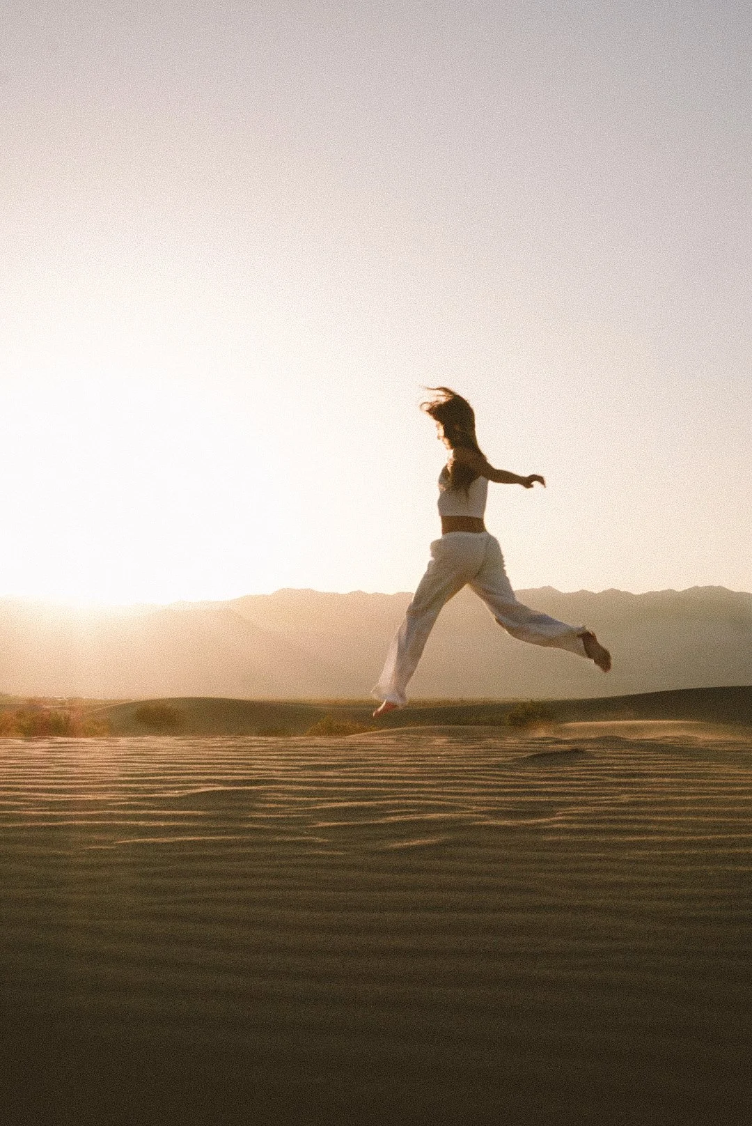 A woman wearing white pants and a white top jumping in the air on a sandy landscape during sunset or sunrise with mountains in the background.