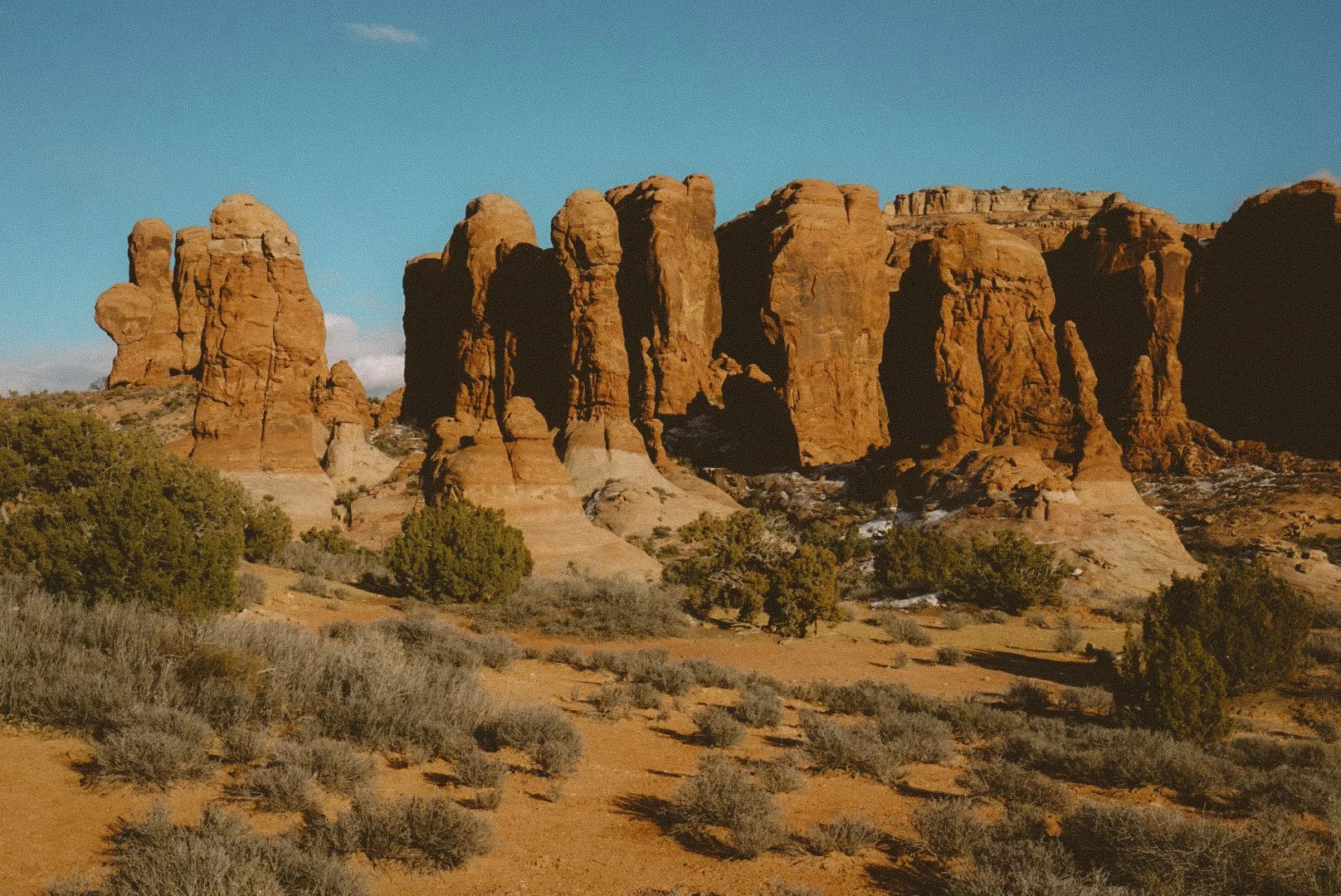 Red rock formations in a desert landscape under a clear blue sky, with sparse bushes and small trees in the foreground.