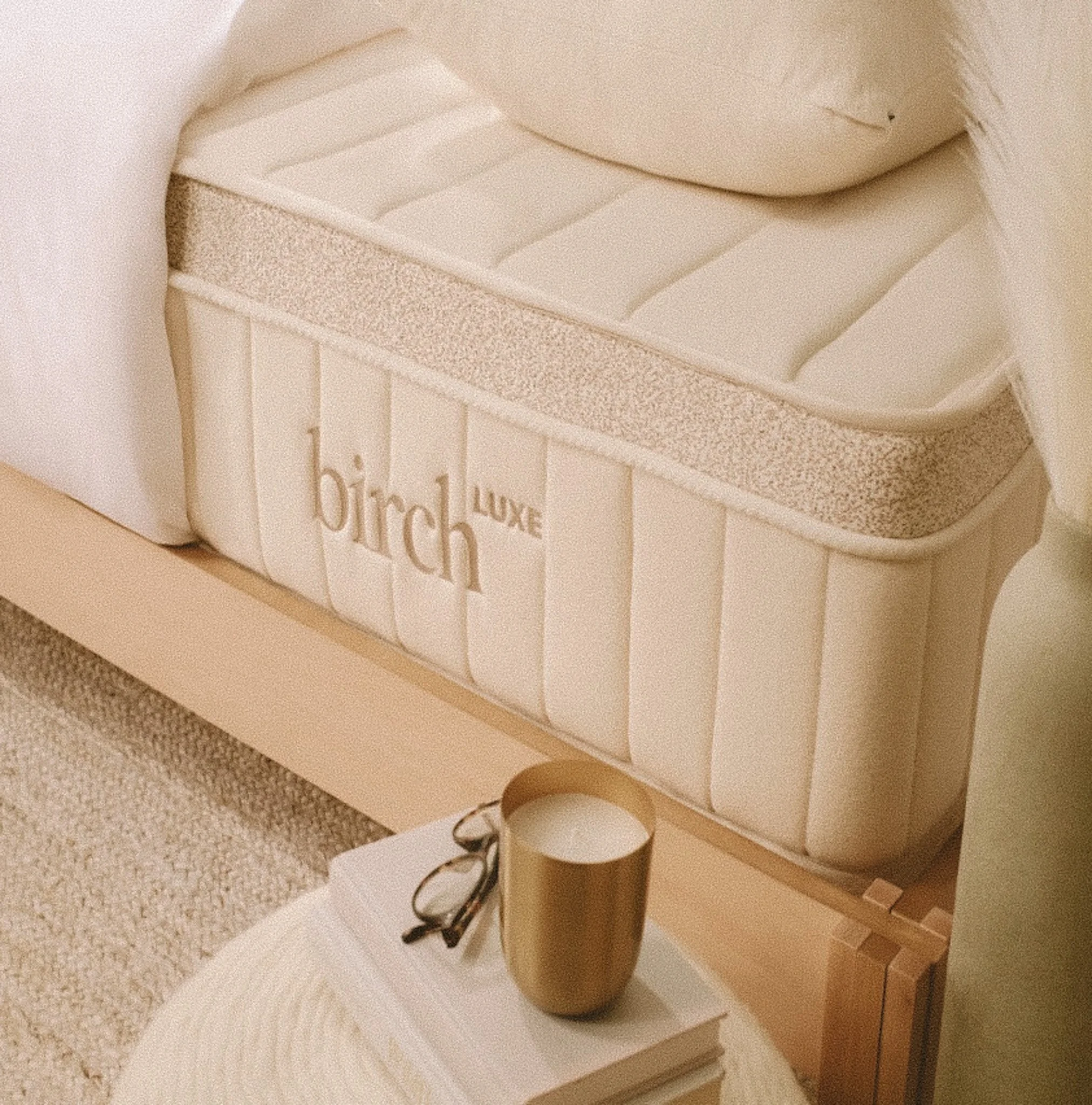 A white mattress with the words 'birch luxe' on the side, a round table with a golden cup, a pair of glasses, and a stack of white books, a plush beige carpet, and a hint of a white sofa.