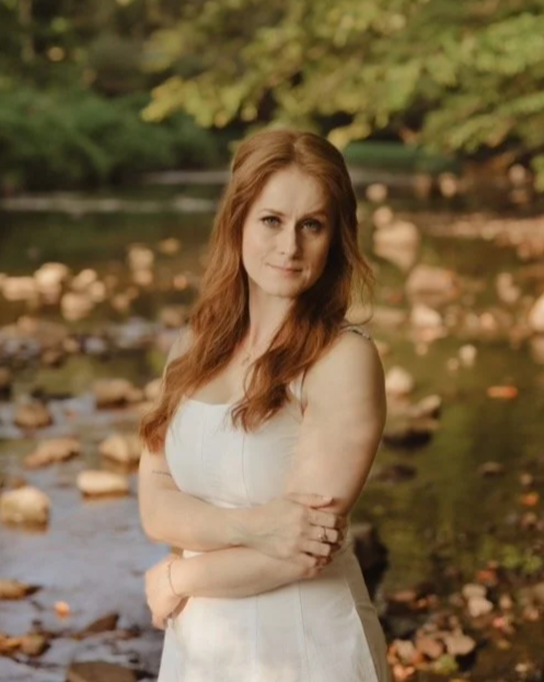 A woman with long red hair and blue eyes smiling, wearing a beige top and gold earrings, against a light textured background.