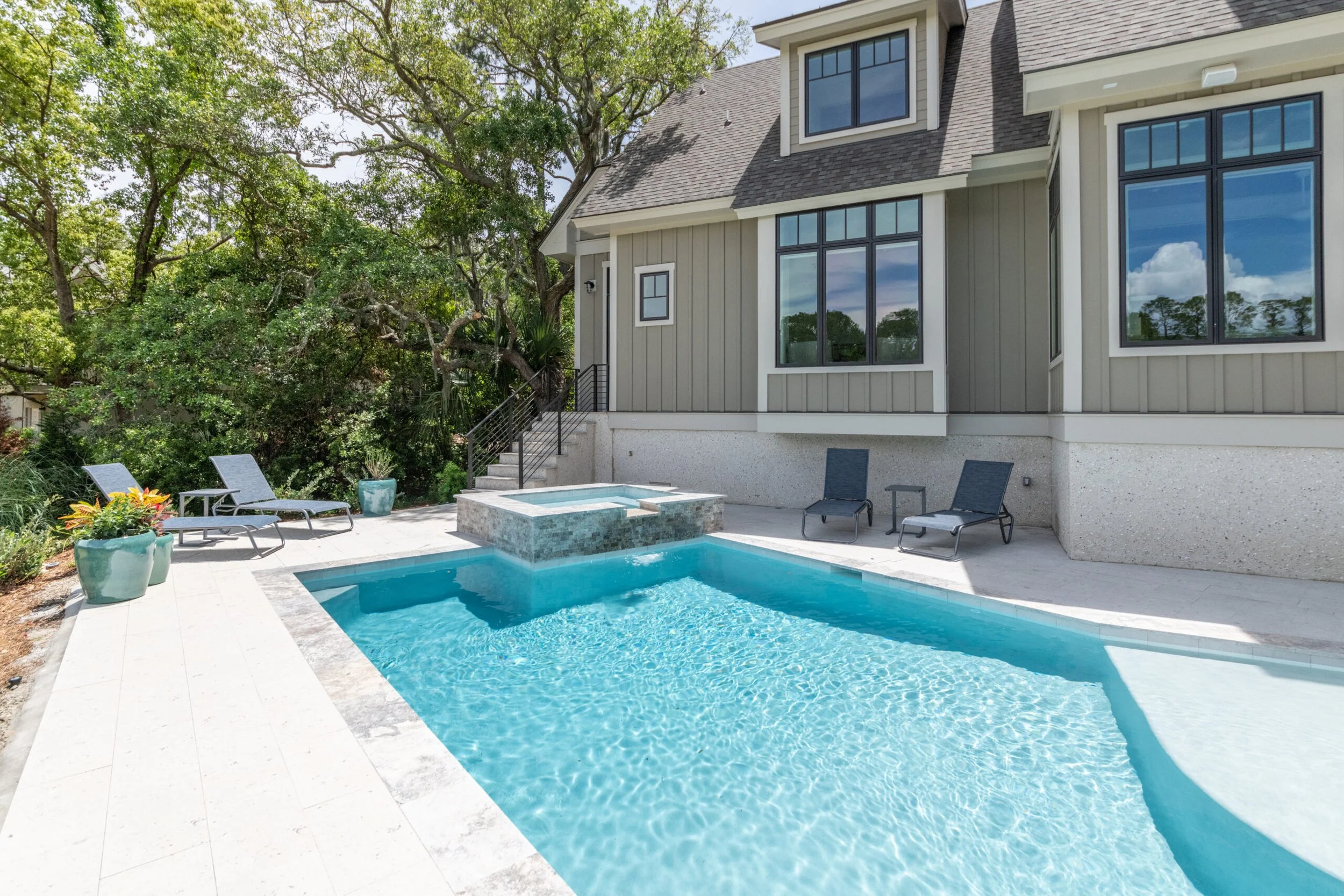 Backyard with a swimming pool and lounge chairs beside a modern house, surrounded by trees. The pool area includes a hot tub with stone accents.