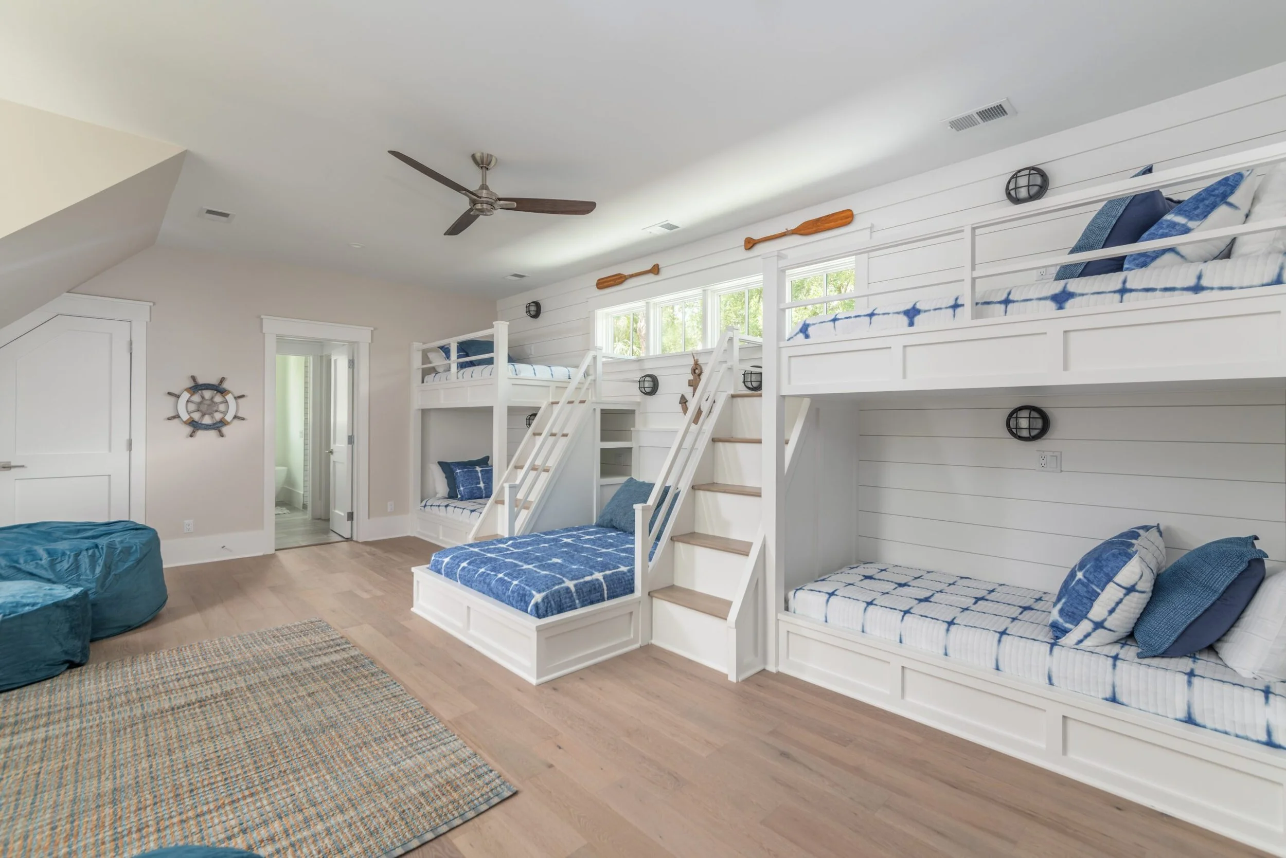 Spacious bedroom with multiple white bunk beds, blue and white bedding, and a nautical theme. Features a wooden floor, ceiling fan, and a nautical wheel decor. Two bean bag chairs and a rug are on the floor, with a view into a bathroom.