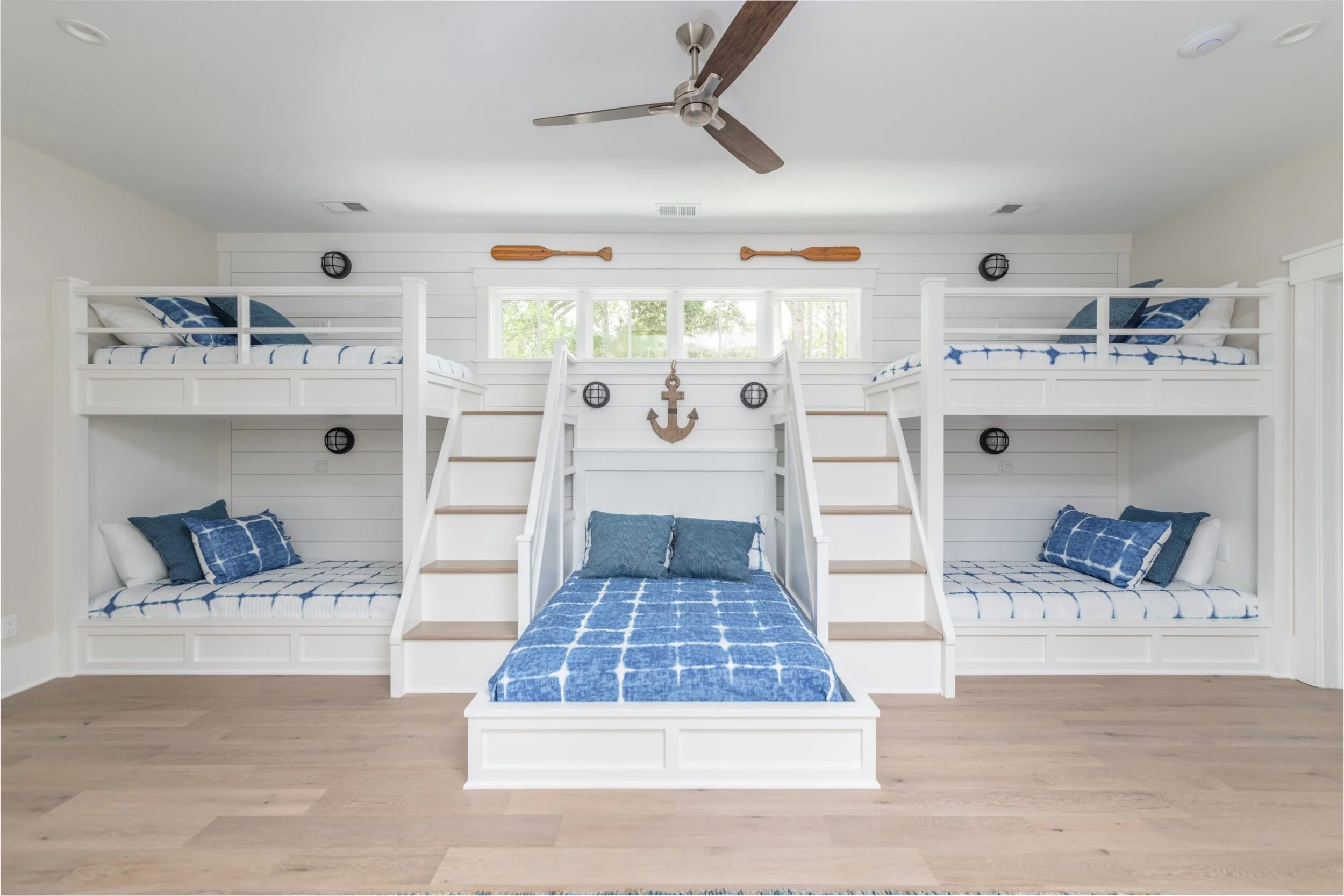 Nautical-themed bedroom with four bunk beds, blue and white bedding, two sets of steps leading to upper beds, wooden paddle decorations, and a wooden anchor on the wall.