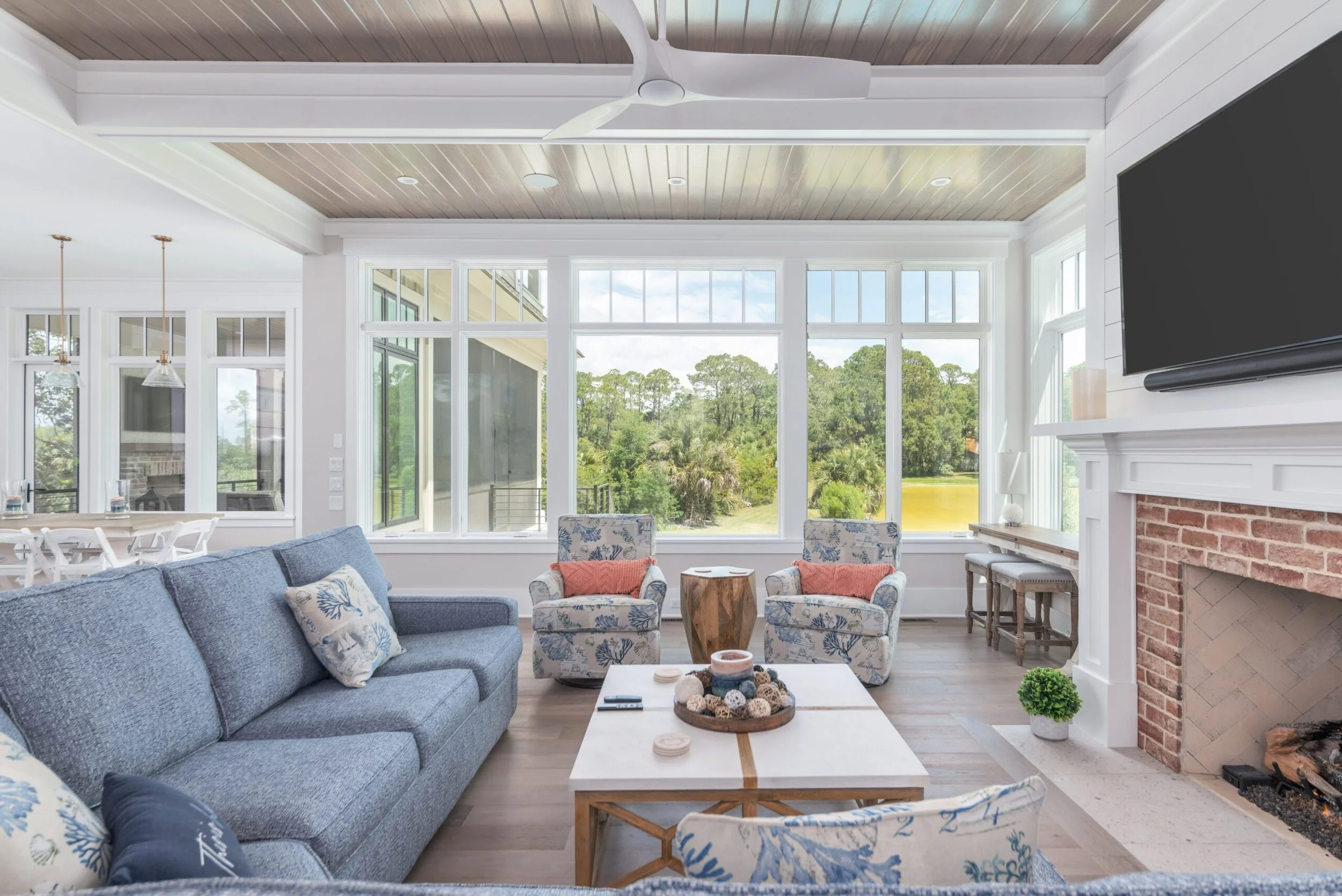 Spacious living room with large windows overlooking a garden, featuring a blue sofa, patterned armchairs with coral cushions, a wooden coffee table, fireplace with TV above, and ceiling fan.