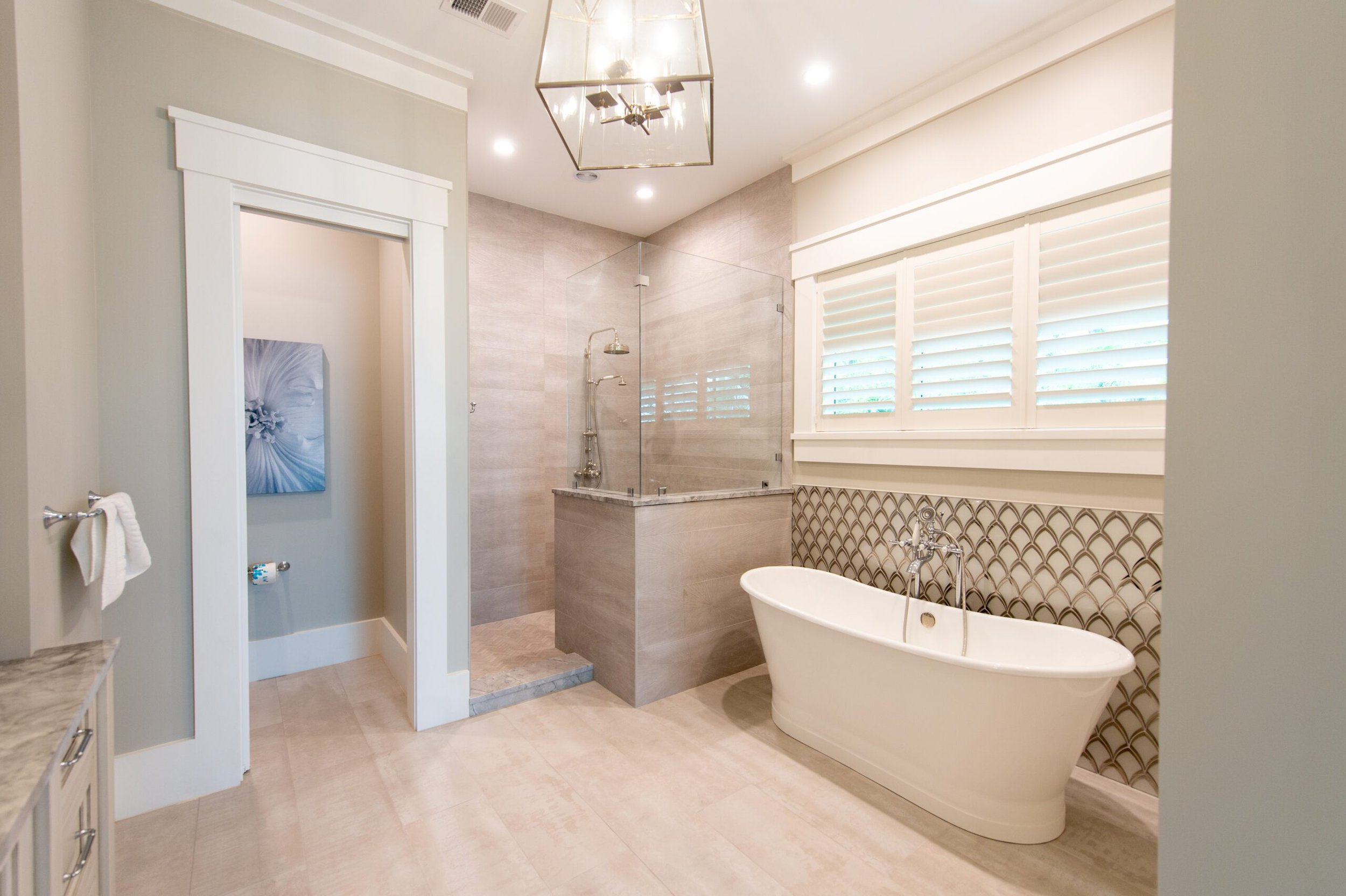 Luxurious modern bathroom with freestanding bathtub, glass-enclosed shower, decorative wall tiles, large window with shutters, and ceiling light fixture.