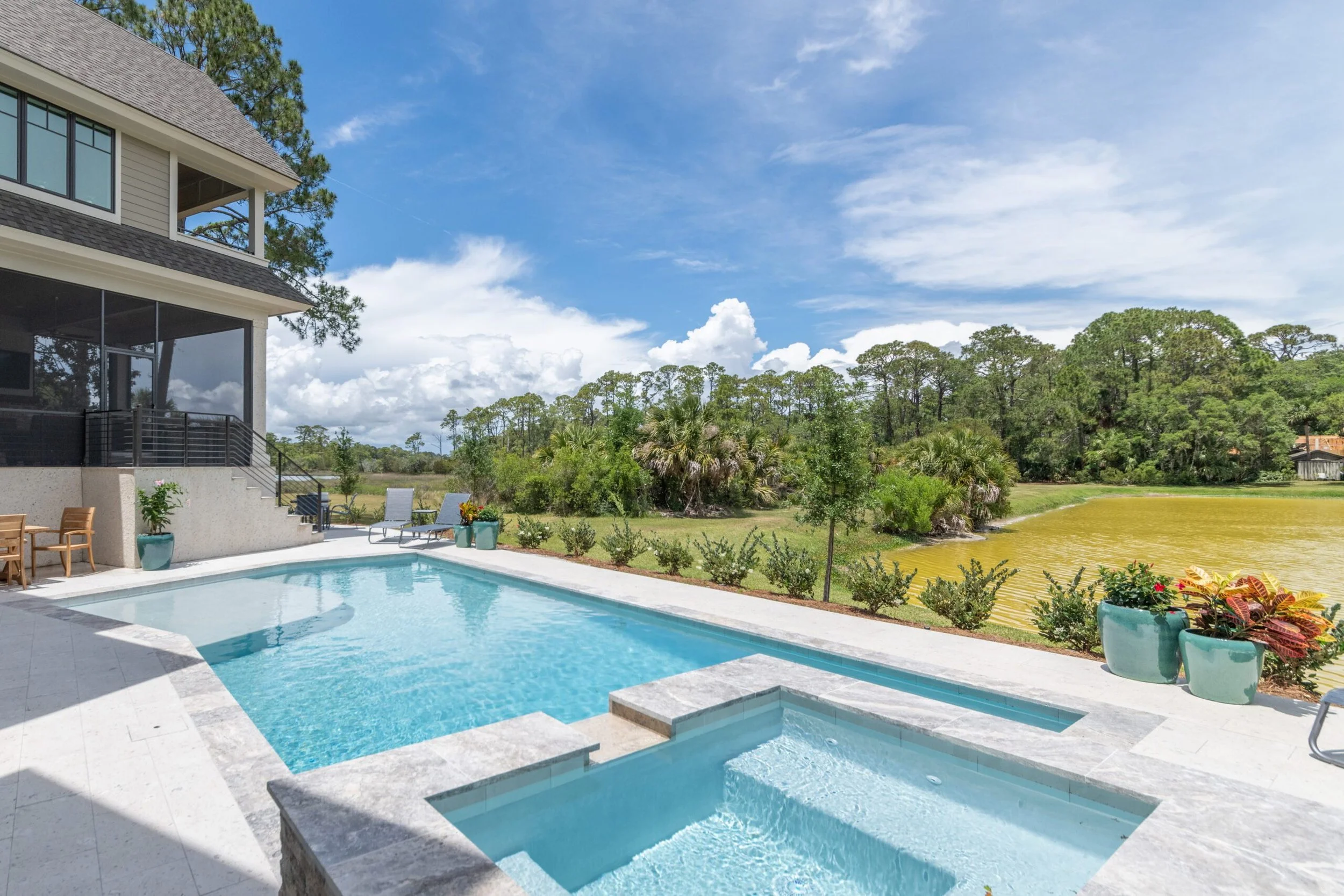 Luxury backyard with a swimming pool, hot tub, and patio furniture, overlooking a scenic lake and lush trees under a blue sky.