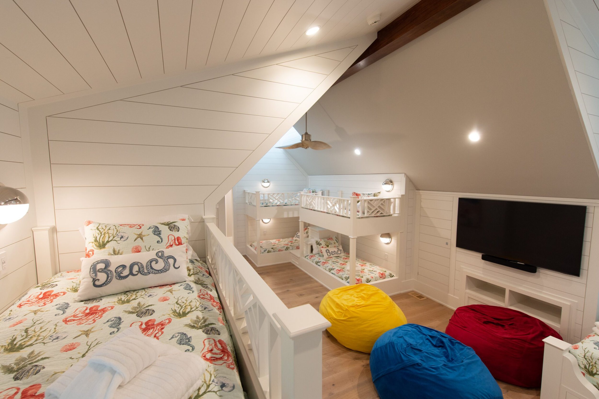 Attic bedroom with three sets of bunk beds, decorated with beach-themed bedding, wooden floors, and colorful bean bags. TV mounted on the wall.