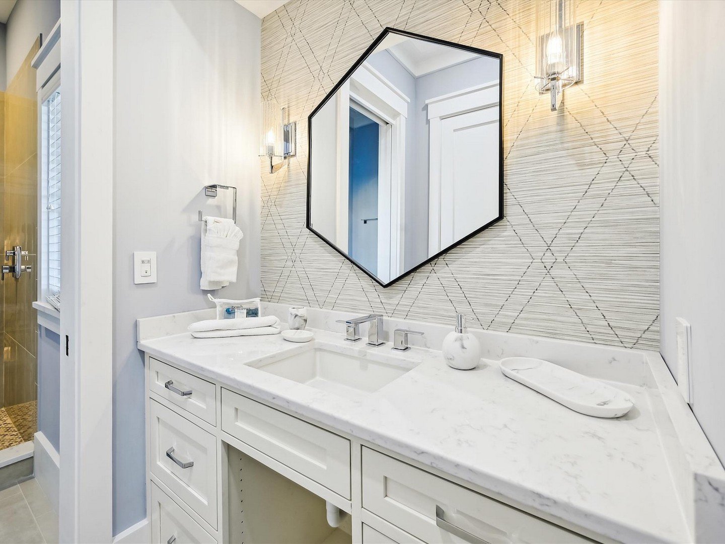 Modern bathroom with white vanity, two sinks, hexagonal mirror, decorative wallpaper, and chrome fixtures