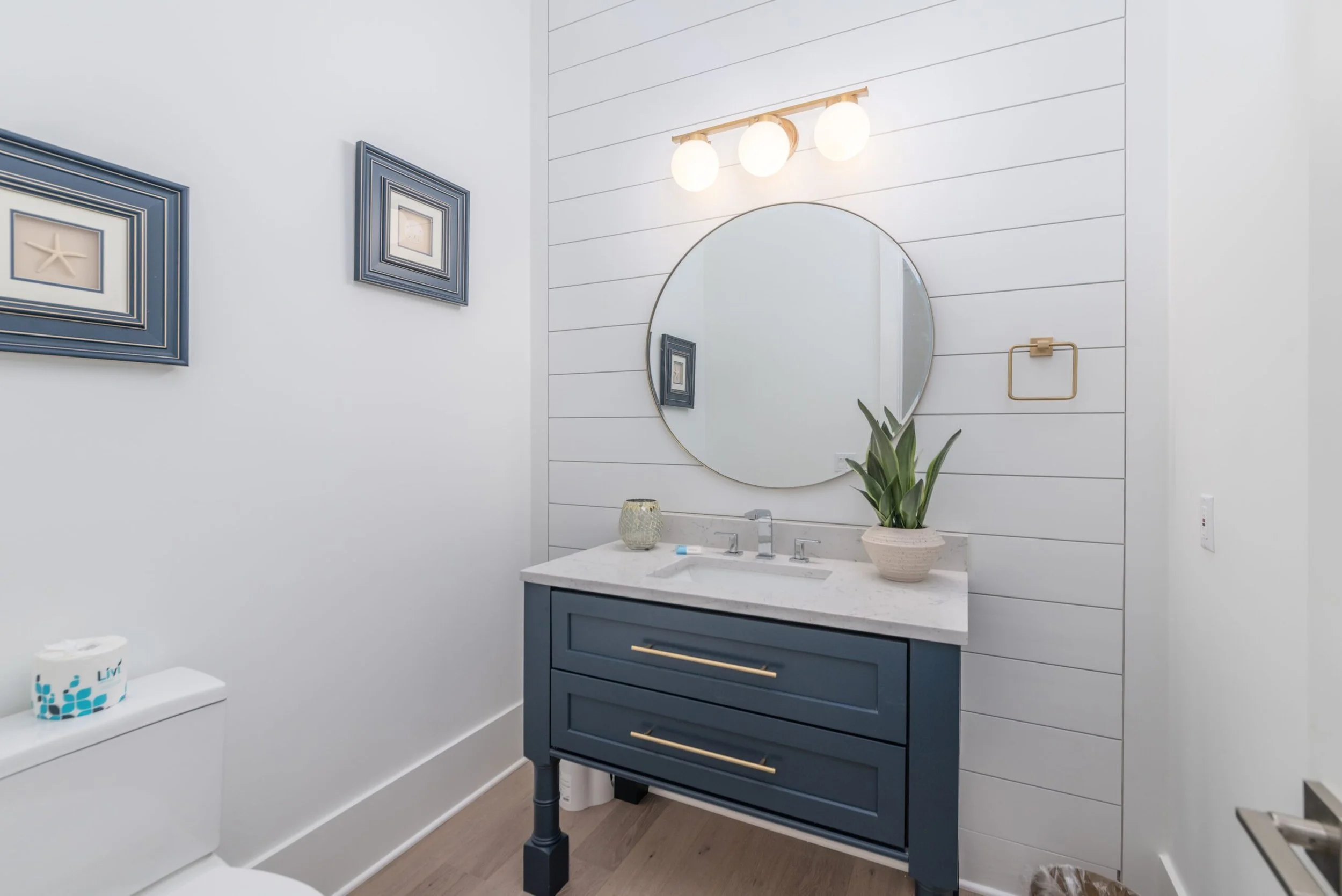 Modern bathroom with a blue vanity, white shiplap walls, round mirror, and three-bulb wall sconce. A potted plant sits on the countertop beside a soap dispenser. The room also has framed starfish art and a roll of toilet paper on the toilet tank.