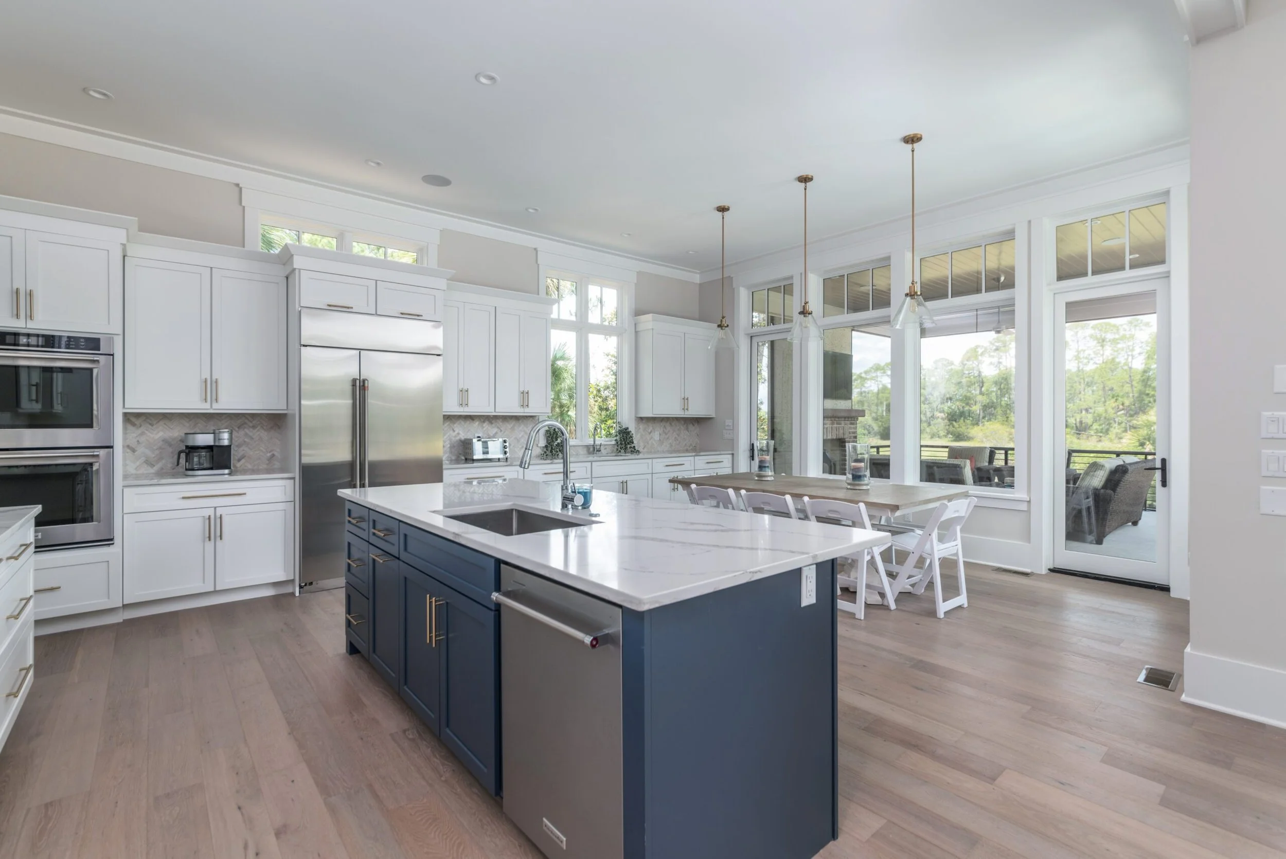 Modern kitchen with white cabinets, stainless steel appliances, large island with sink, pendant lights, hardwood floors, and a view of the outdoors.