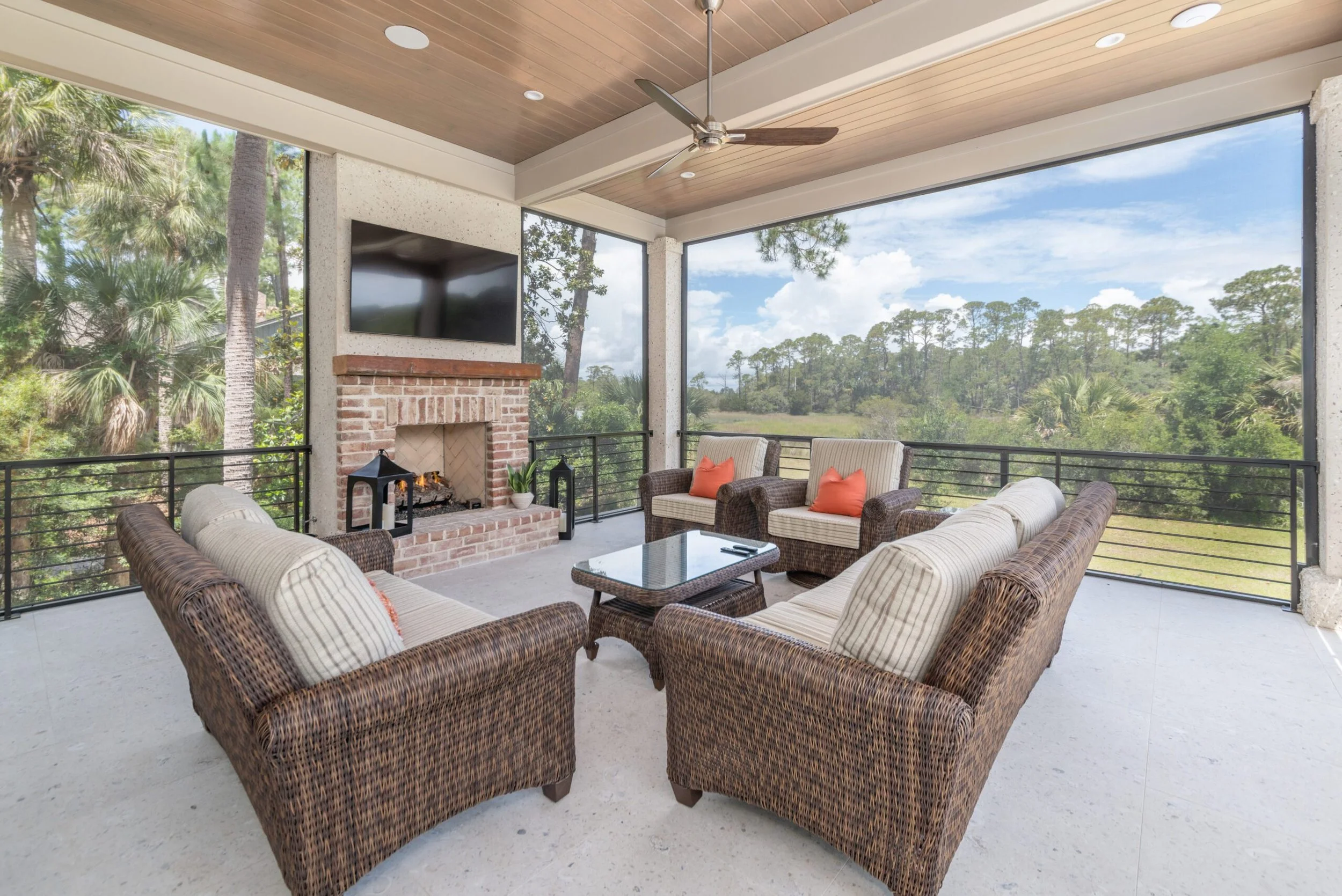 Outdoor patio with fireplace, wicker furniture, ceiling fan, wall-mounted TV, surrounded by nature views.