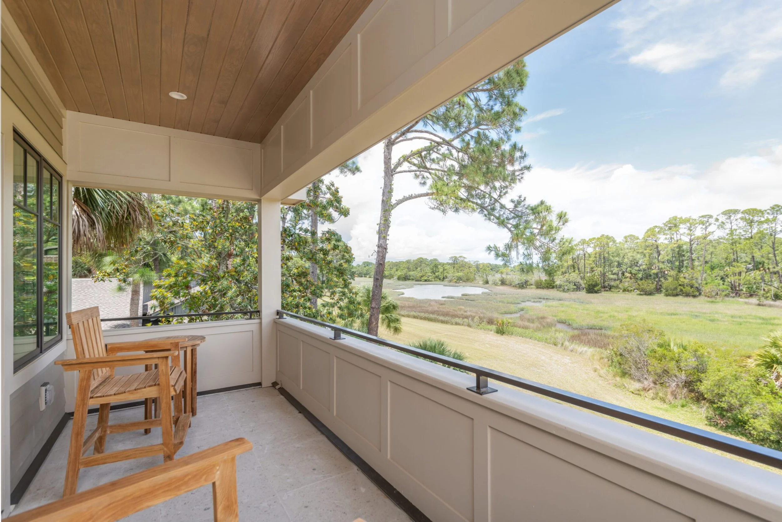 Balcony view with wooden chairs overlooking a green landscape and a small pond, surrounded by trees under a clear blue sky.