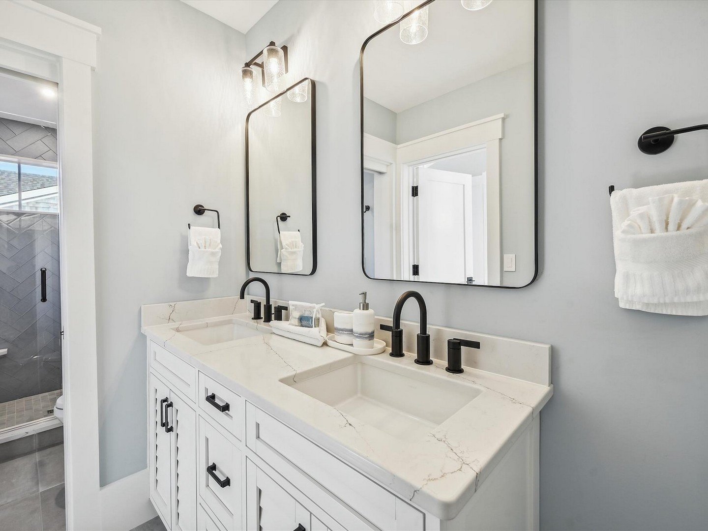 Modern bathroom with dual mirrors, marble countertop, black faucets, and white cabinetry. Features a towel rack with folded towels and a visible shower area with herringbone tile design.