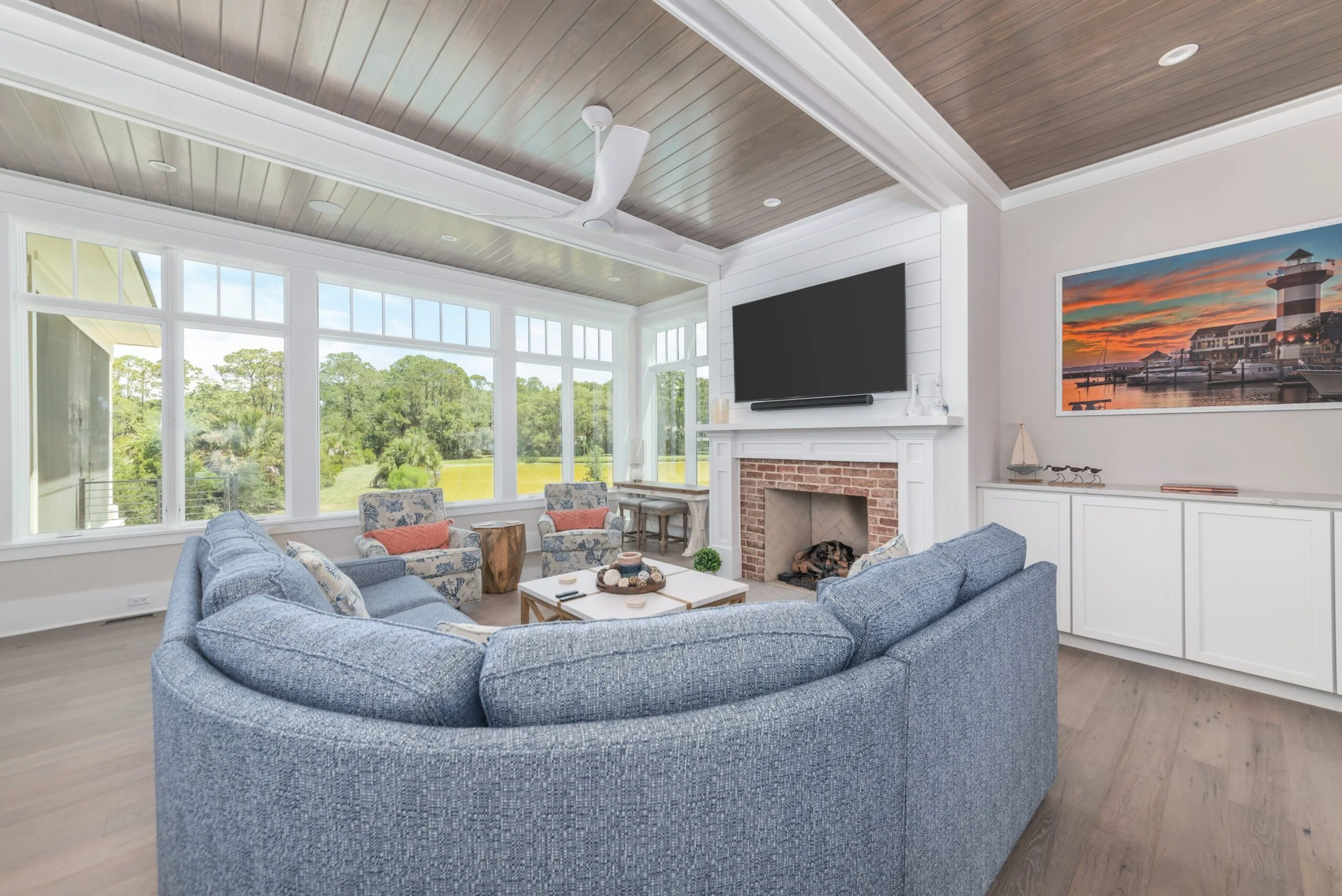 Spacious living room with blue sectional sofa, large windows, flat-screen TV above fireplace, and coastal-themed decor.