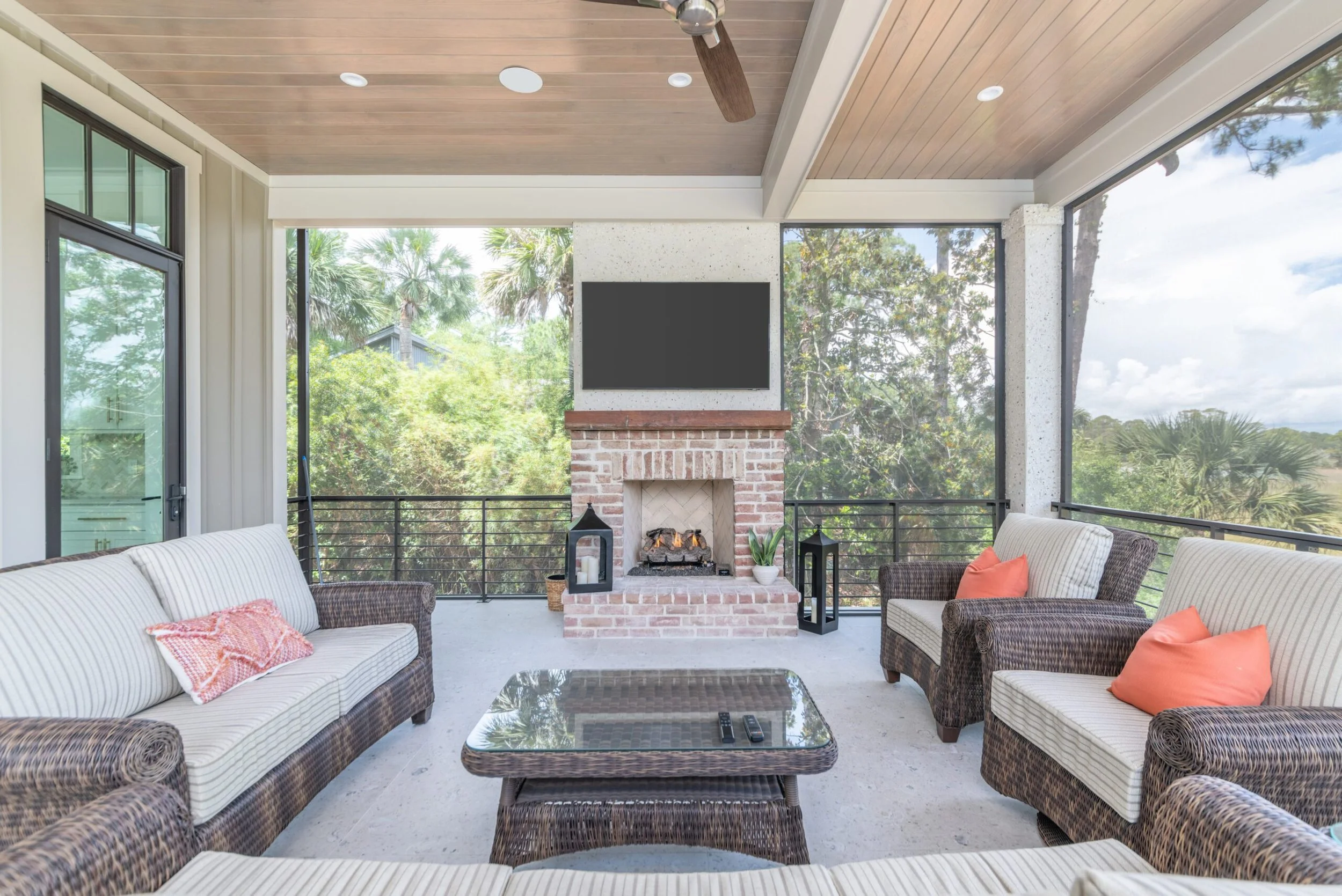 Outdoor screened-in patio with wicker furniture, striped cushions, and orange pillows. Features a brick fireplace with a TV above it, ceiling fan, and views of lush greenery.