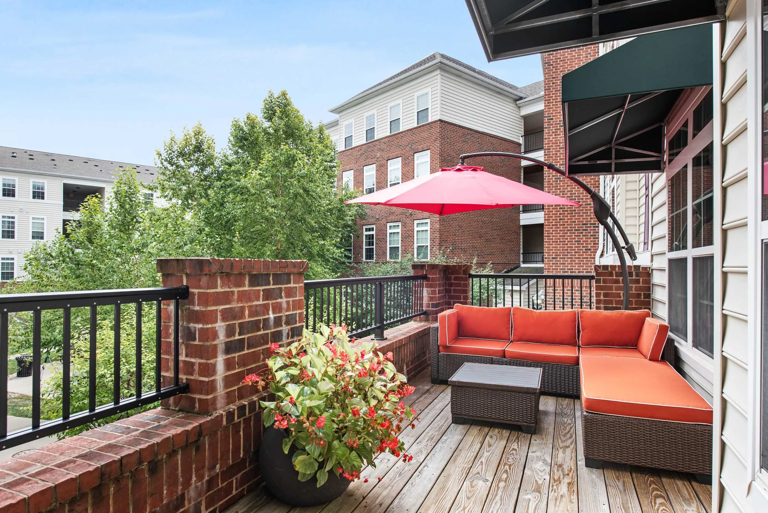Outdoor balcony with a brick railing, red cushioned wicker sectional sofa, a small black table, pink umbrella, potted plant with red flowers, and neighboring brick and white apartment buildings.