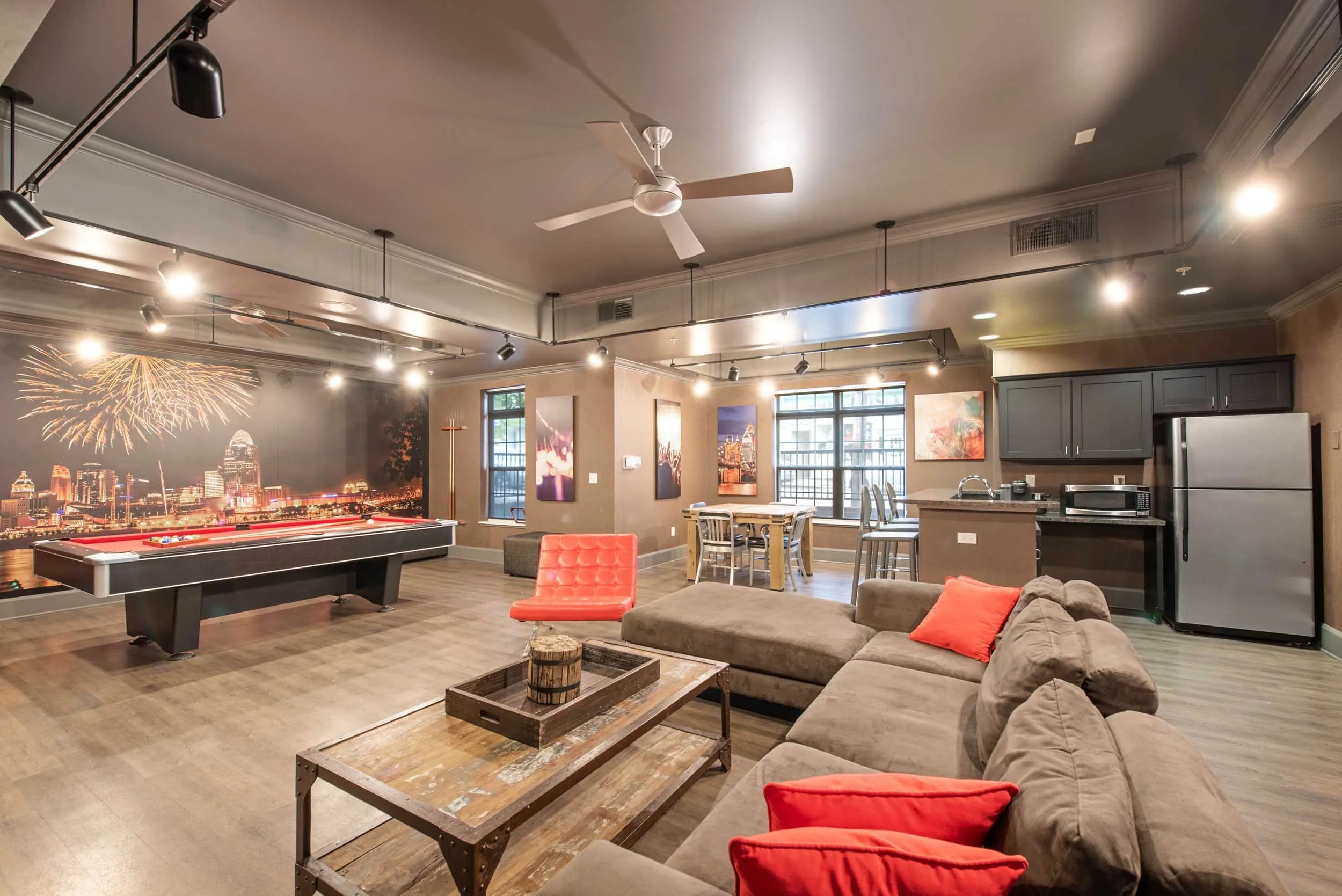 A spacious living room with a pool table, a sectional sofa with red pillows, a wooden coffee table, and a kitchen area in the background. The room has large windows, colorful wall art, and a cityscape mural on one wall with fireworks.