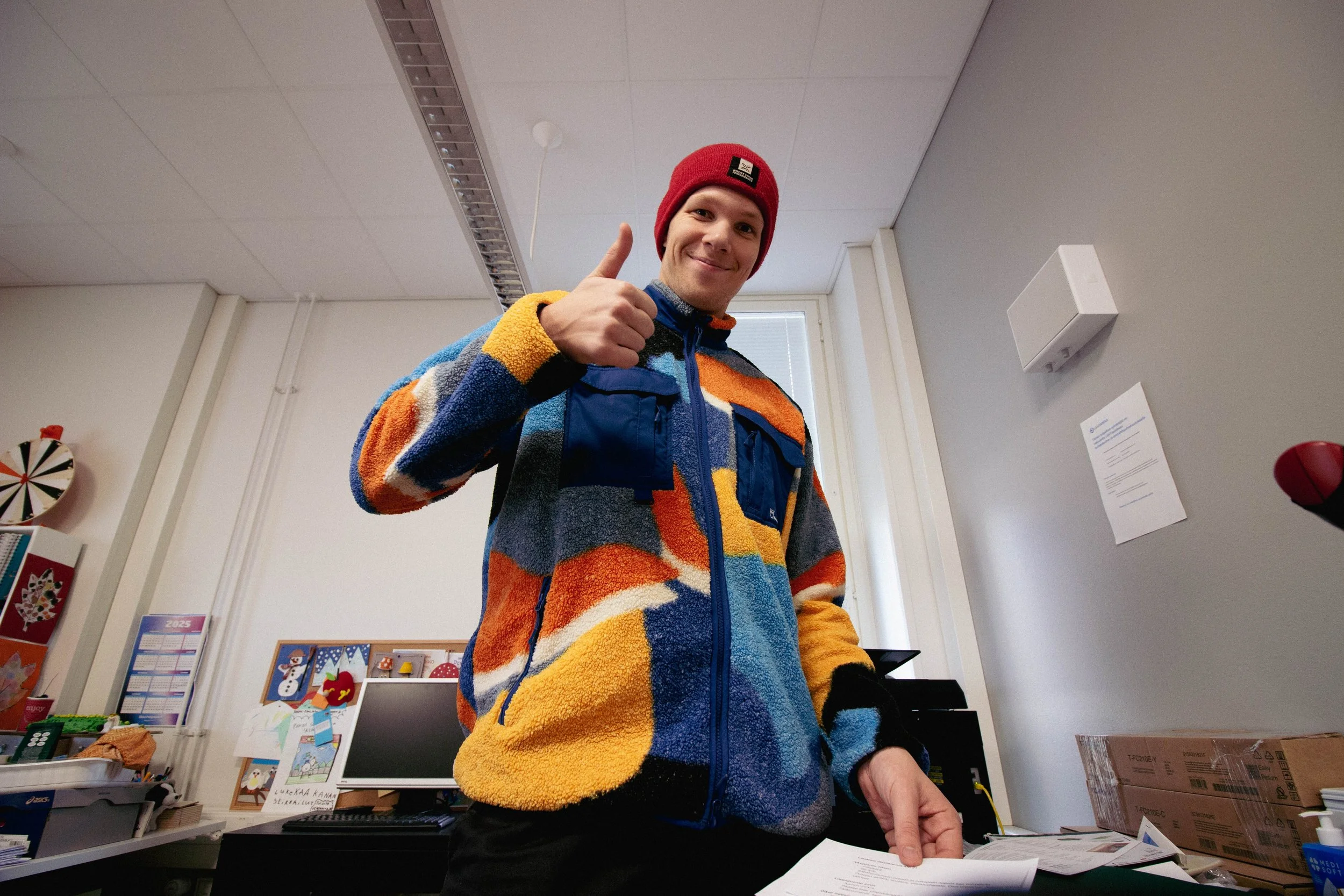 Hymyilevä nuori mies punaisessa beanie-hattussa ja värikkäässä fleecetakissa suuressa sisätilassa, ehkä konttorissa tai koulussa, taustalla valkoseiniä ja ikkuna.