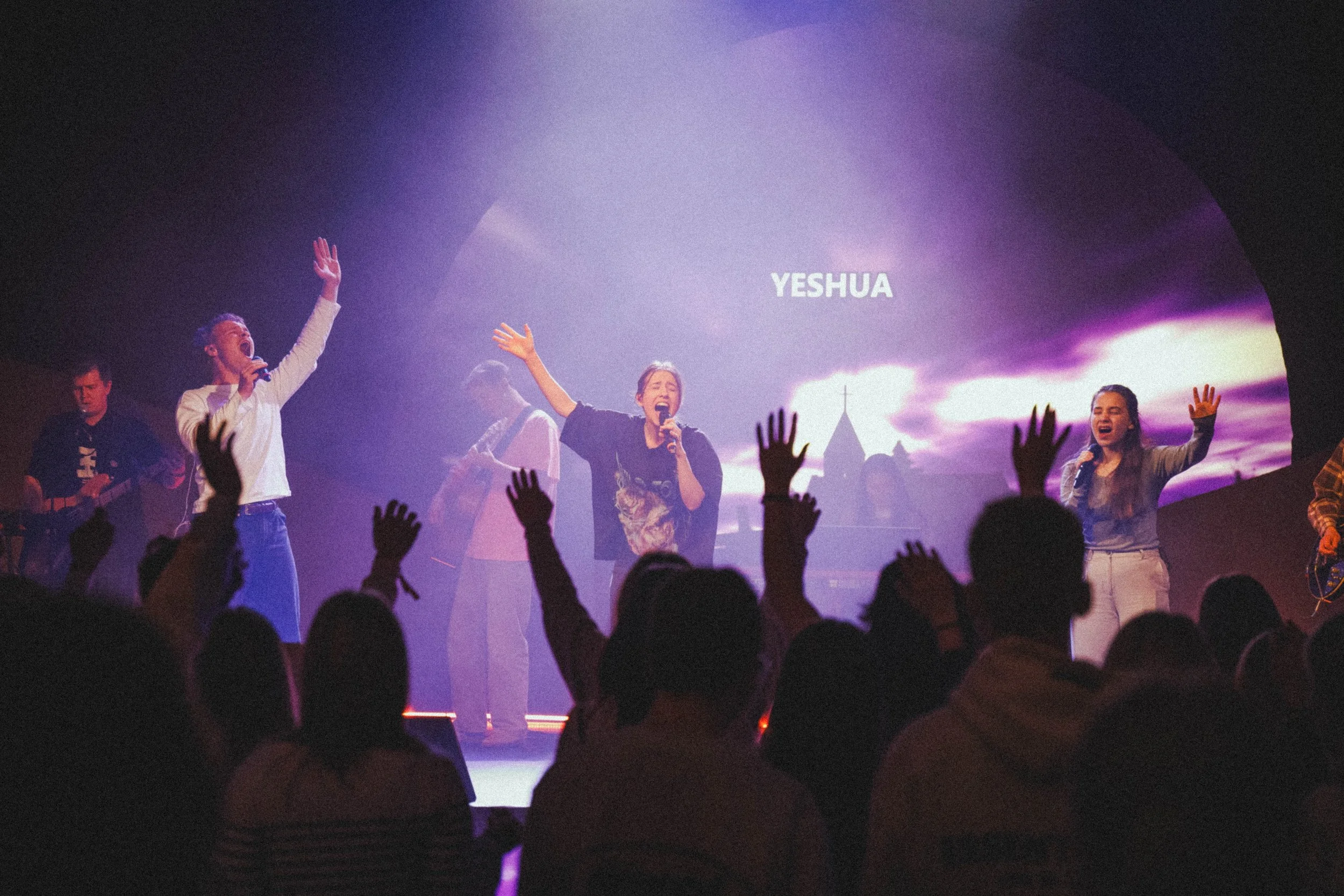 Ryhmä ihmisiä ylistää Jumalaa tapahtumassa, jossa näkyy teksti 'Yeshua' taustalla.