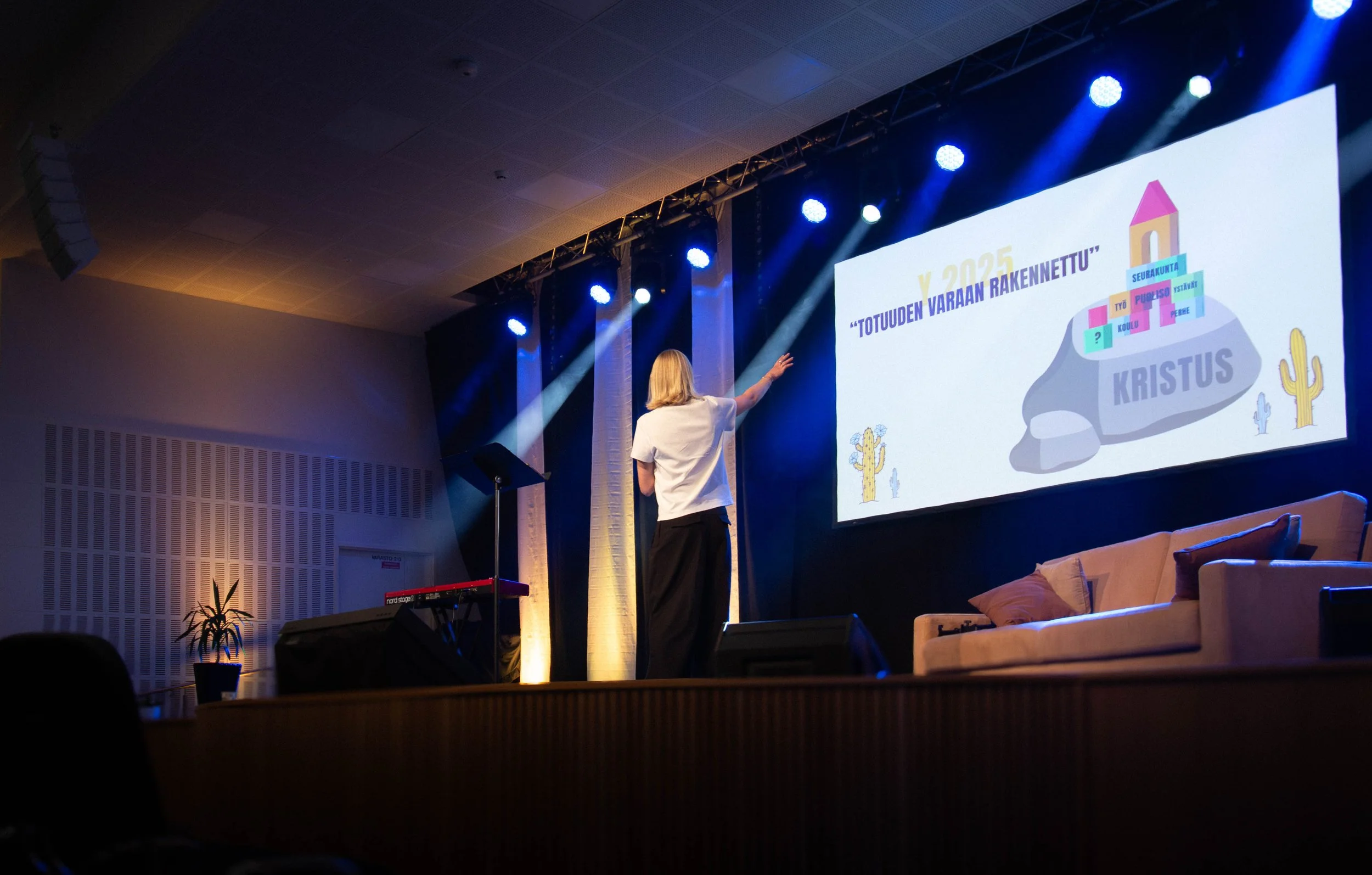 Henkilö esittää kuvaa, jossa on teatterinäyttö suurella ruudulla, jonka kuva sisältää rakennusteemaisen kuvituksen ja tekstejä. Henkilö on katsomassa tai esittelemässä sisältöä.