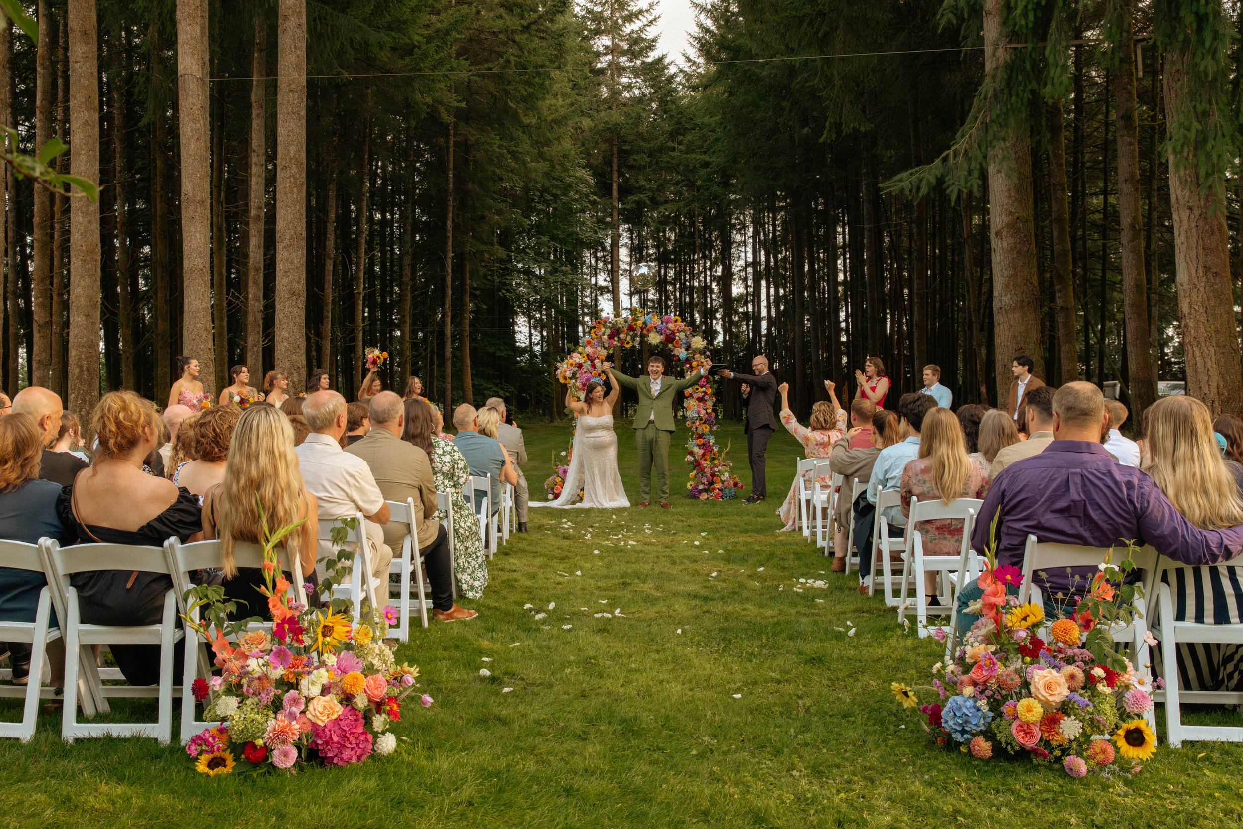A wedding ceremony takes place outdoors in a forest clearing. The bride and groom are at the altar with a floral arch behind them, surrounded by guests seated on white chairs. Floral arrangements are at the end of each row, and the couple is celebrating with smiles.