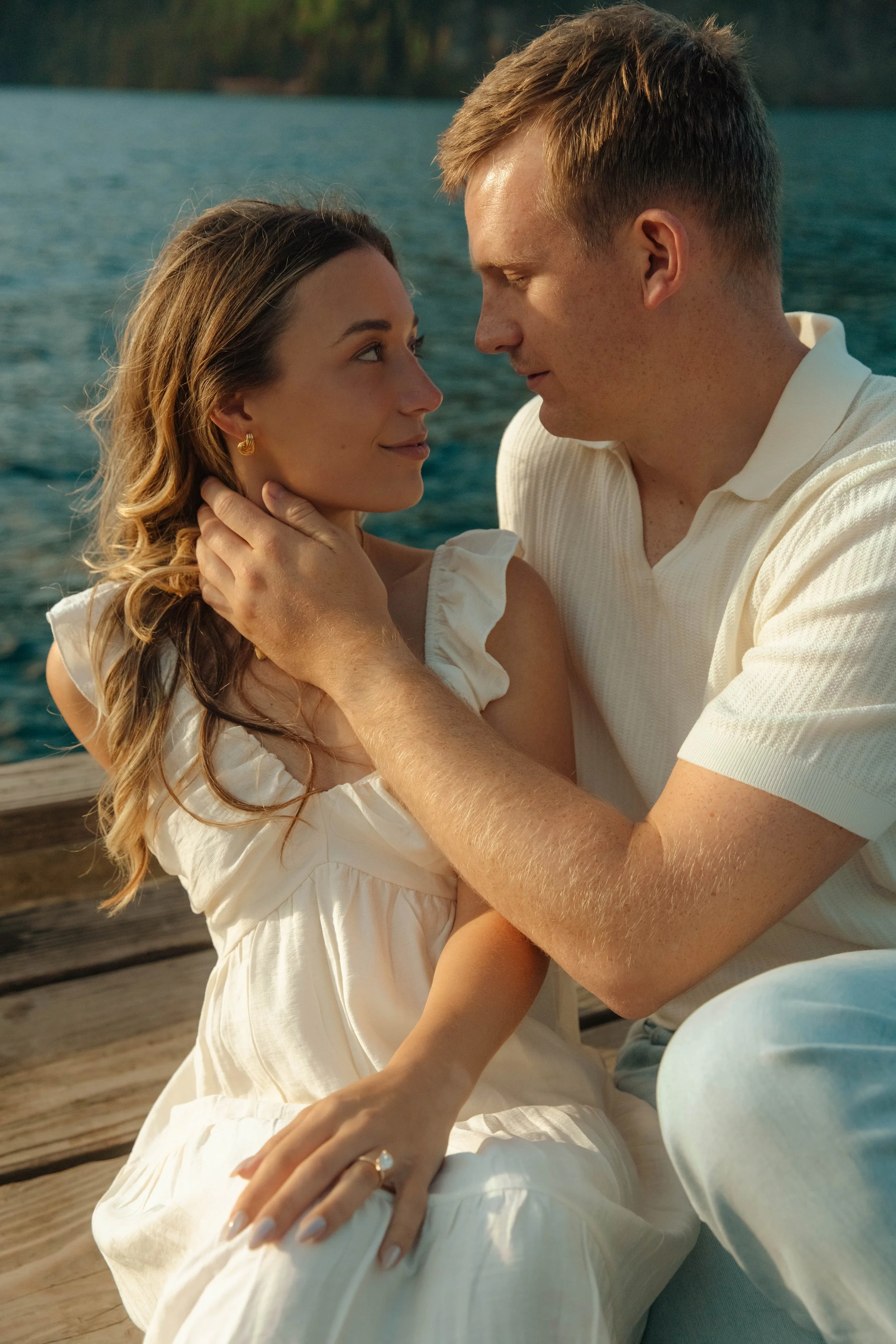 A man and woman sitting on a dock by the water, gazing into each other's eyes in a romantic moment, with the woman wearing a white dress and the man in a cream shirt.