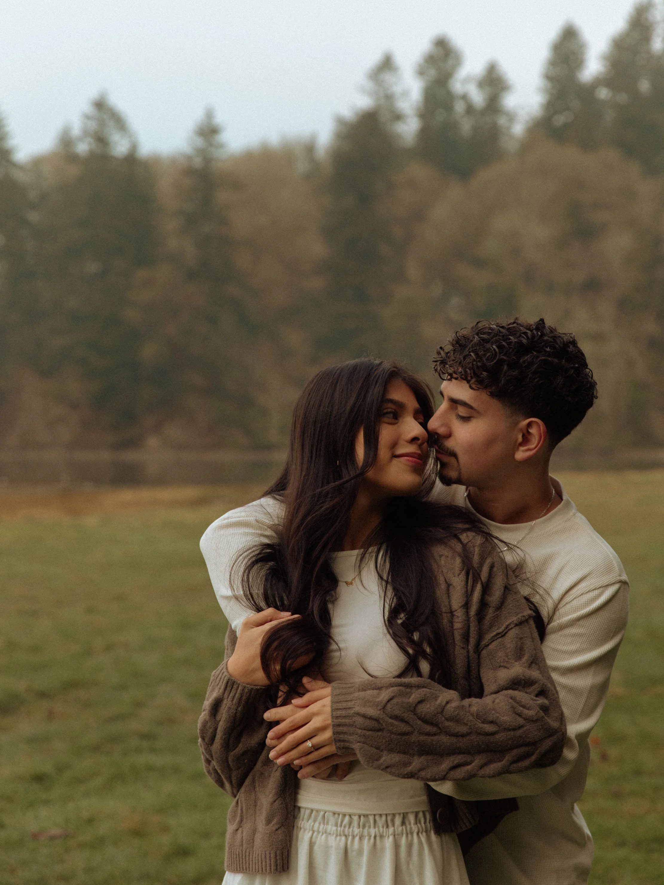 A wedding ceremony takes place outdoors in a forest clearing. The bride and groom are at the altar with a floral arch behind them, surrounded by guests seated on white chairs. Floral arrangements are at the end of each row, and the couple is celebrating with smiles.