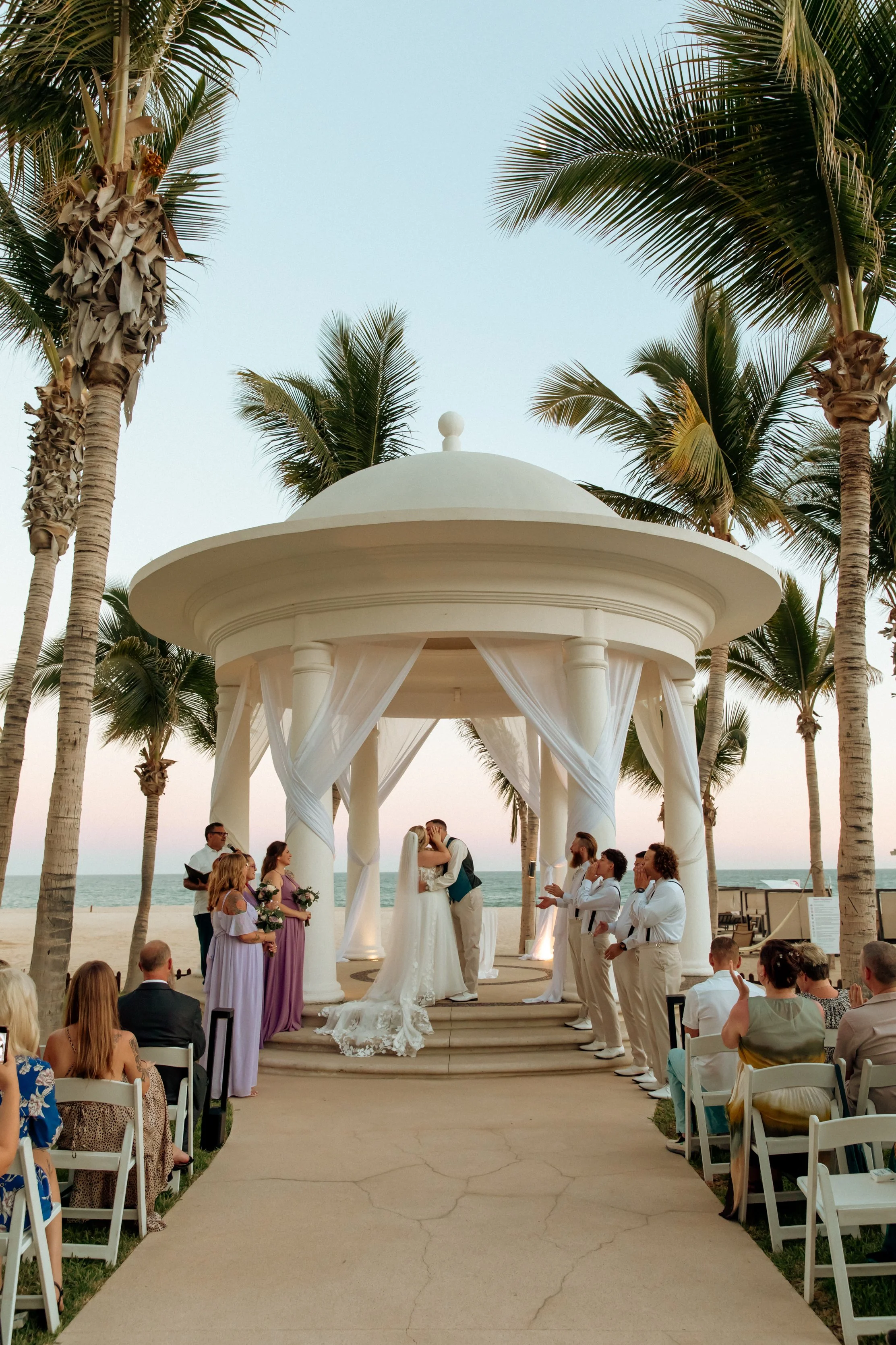 A wedding ceremony taking place under a white gazebo on the beach, with the ocean and palm trees in the background, during sunset.