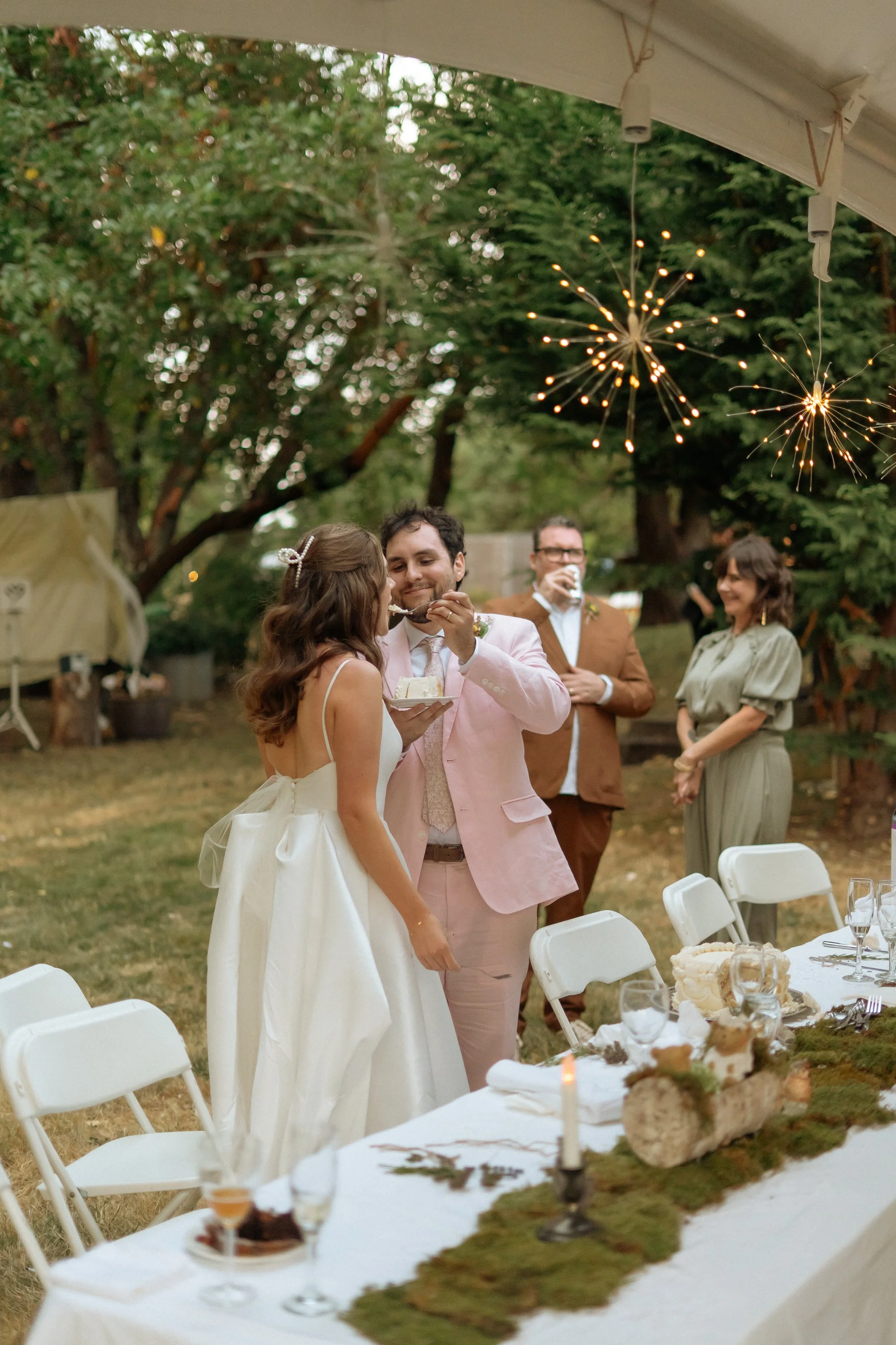 Wedding celebration outdoors with a bride and groom feeding each other cake, surrounded by friends, under string lights and sparklers, with decorated table and lush green trees.