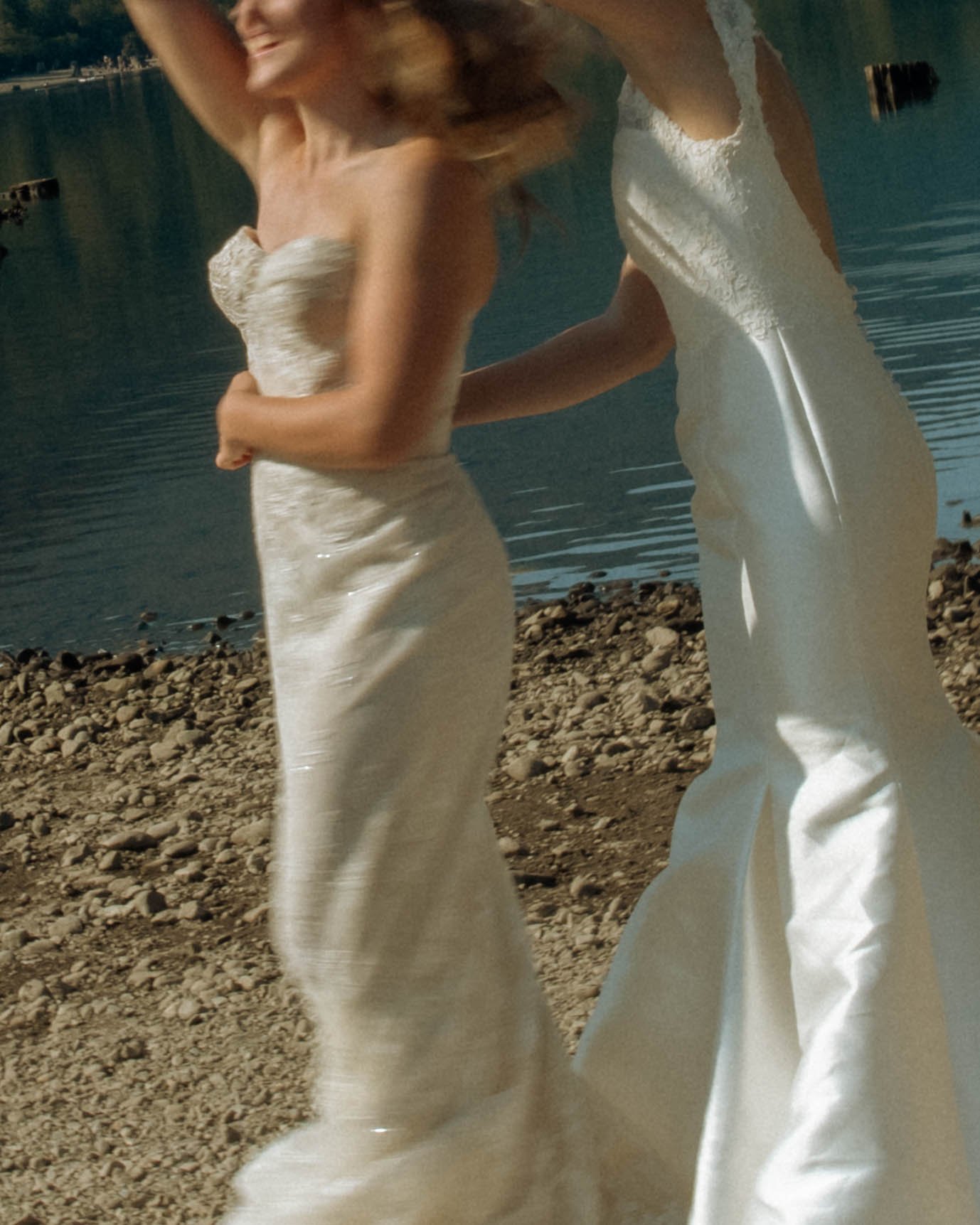 Two women in white dresses standing on a rocky lakeshore, smiling and embracing each other.