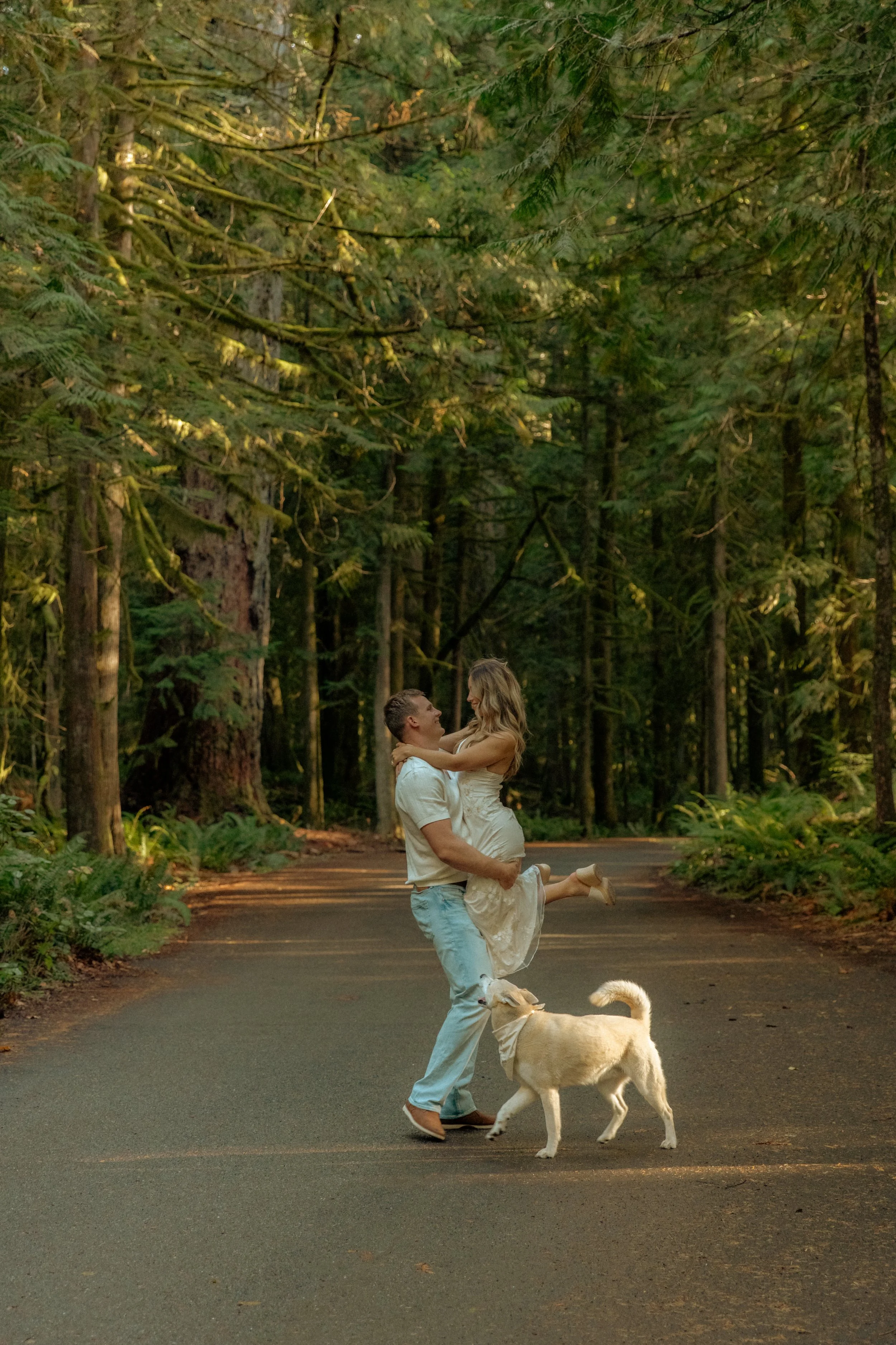 A couple, with the man lifting the woman and both smiling, standing on a forest road with a dog nearby. Dense greenery surrounds them.