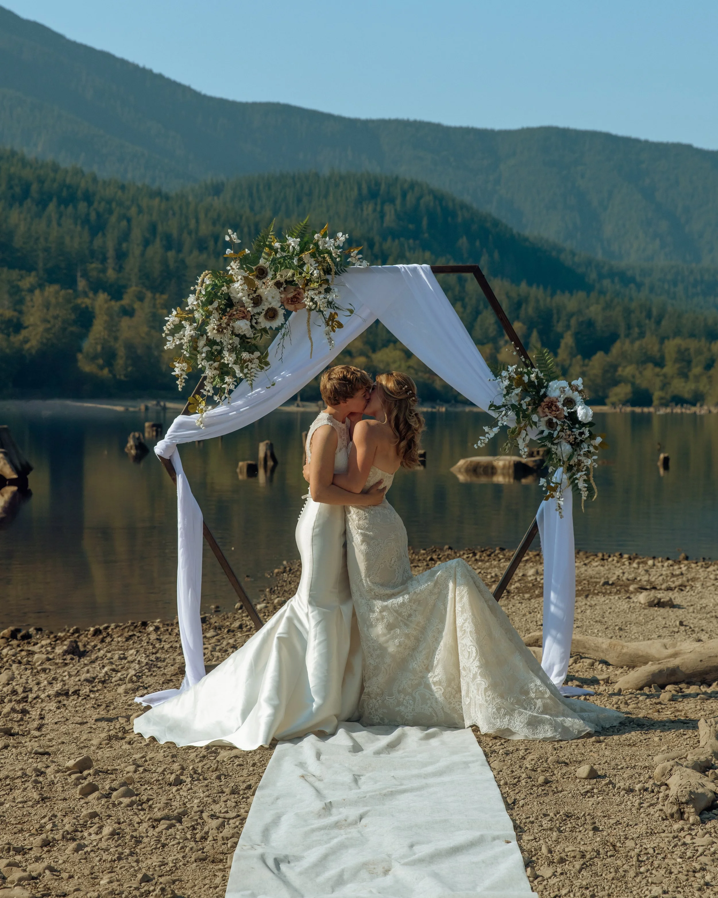 Two women in wedding dresses sharing a kiss under a floral wedding arch on a lakeshore with mountains in the background.