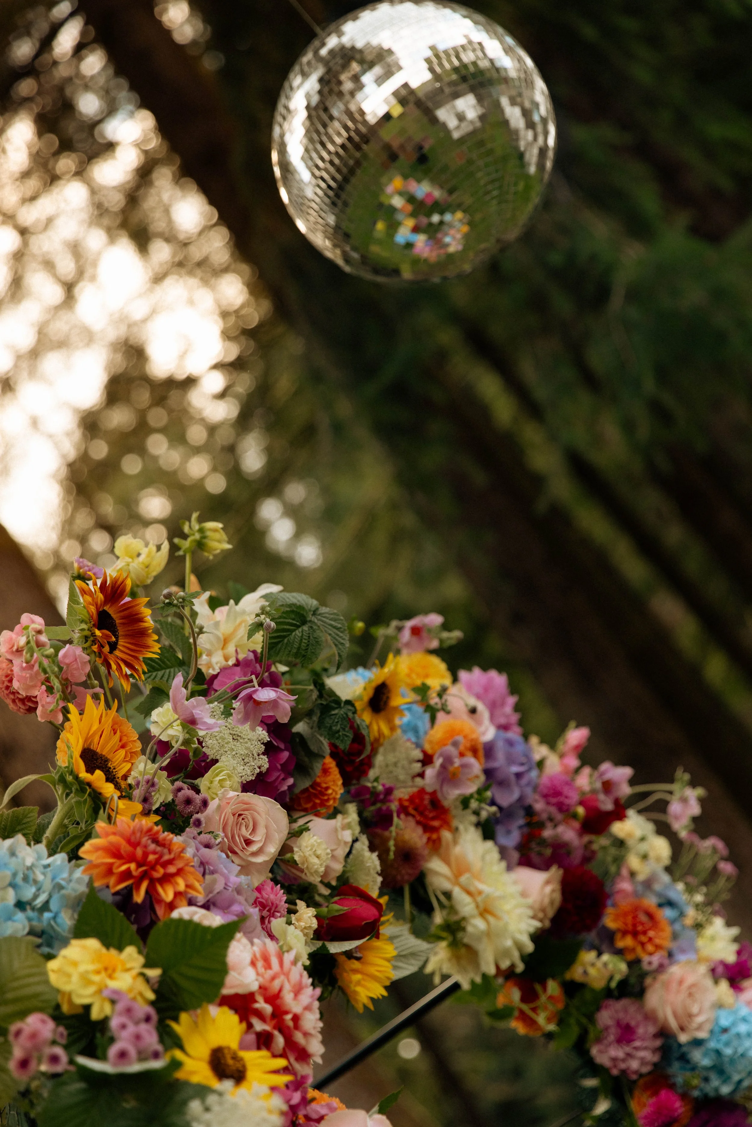 A large bouquet of colorful flowers, including sunflowers, roses, and various other blooms, with a disco ball hanging above outdoors.