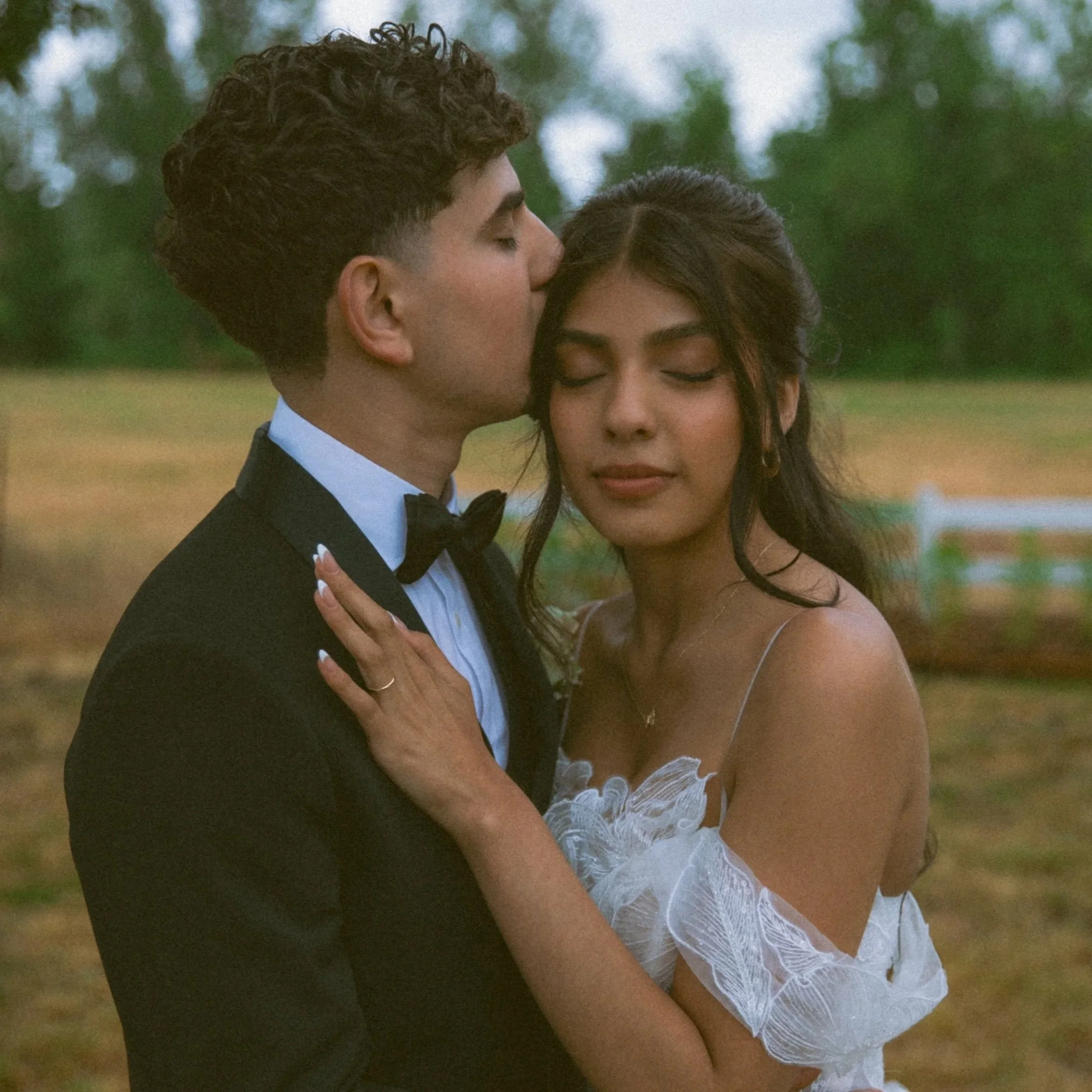 A couple dressed in formal wedding attire, with the groom kissing the bride on her forehead outdoors, with trees and grass in the background.